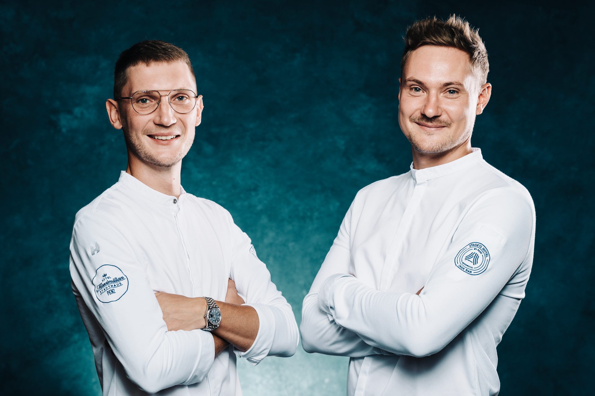 Two smiling men in white uniforms with crossed arms on a blue-green background.