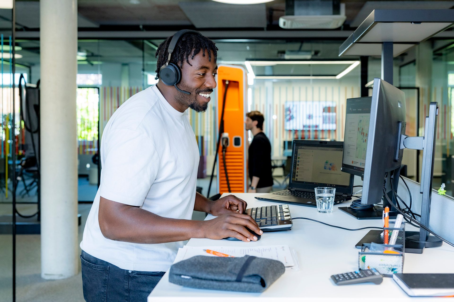 A smiling man wearing headphones works at a desk with a laptop and monitor in a modern office.