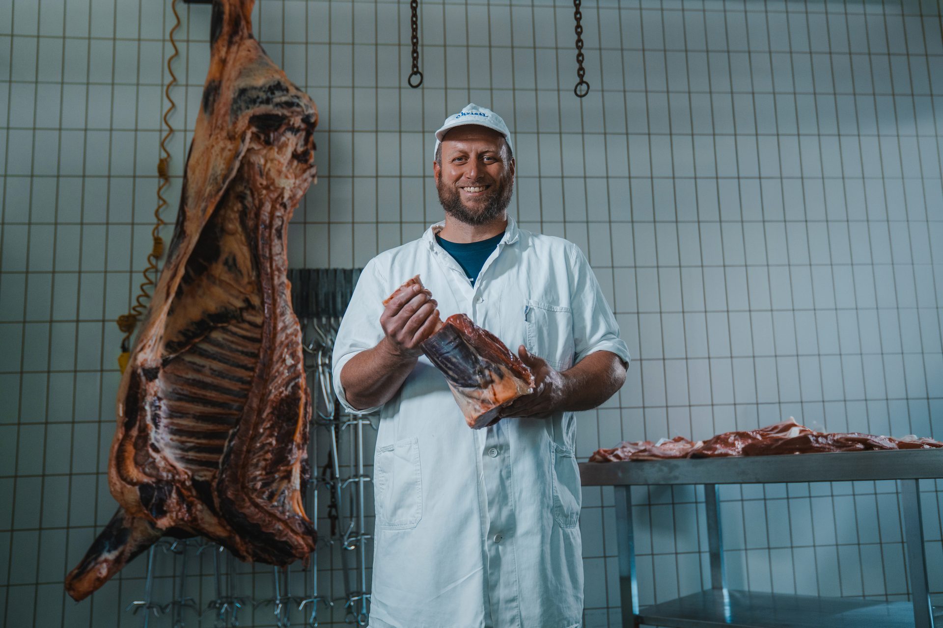 A smiling butcher holds a cut of meat in a processing room with a hanging carcass and more meat.
