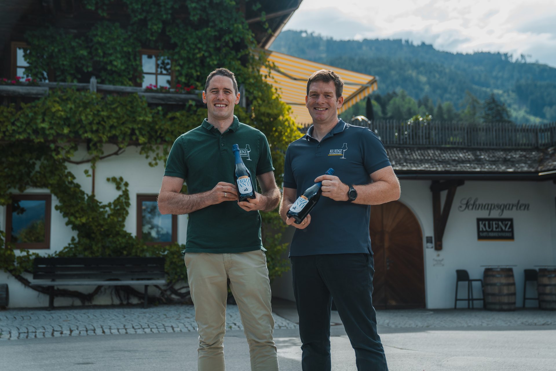Two men holding bottles, smiling in front of Schnapsgarten Kuenz with mountains.