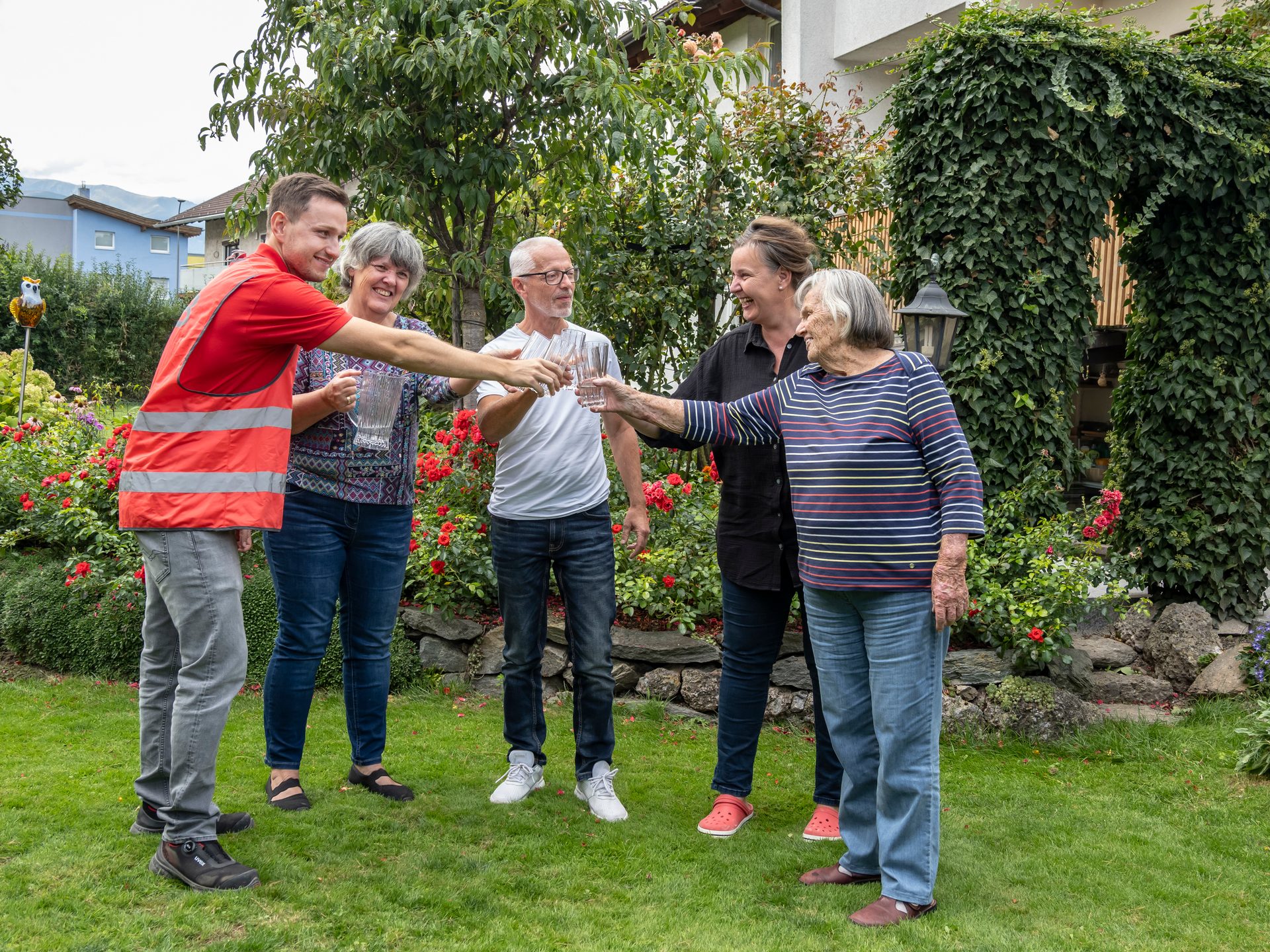 Five people of different ages cheerfully toasting with water glasses in a lush garden.