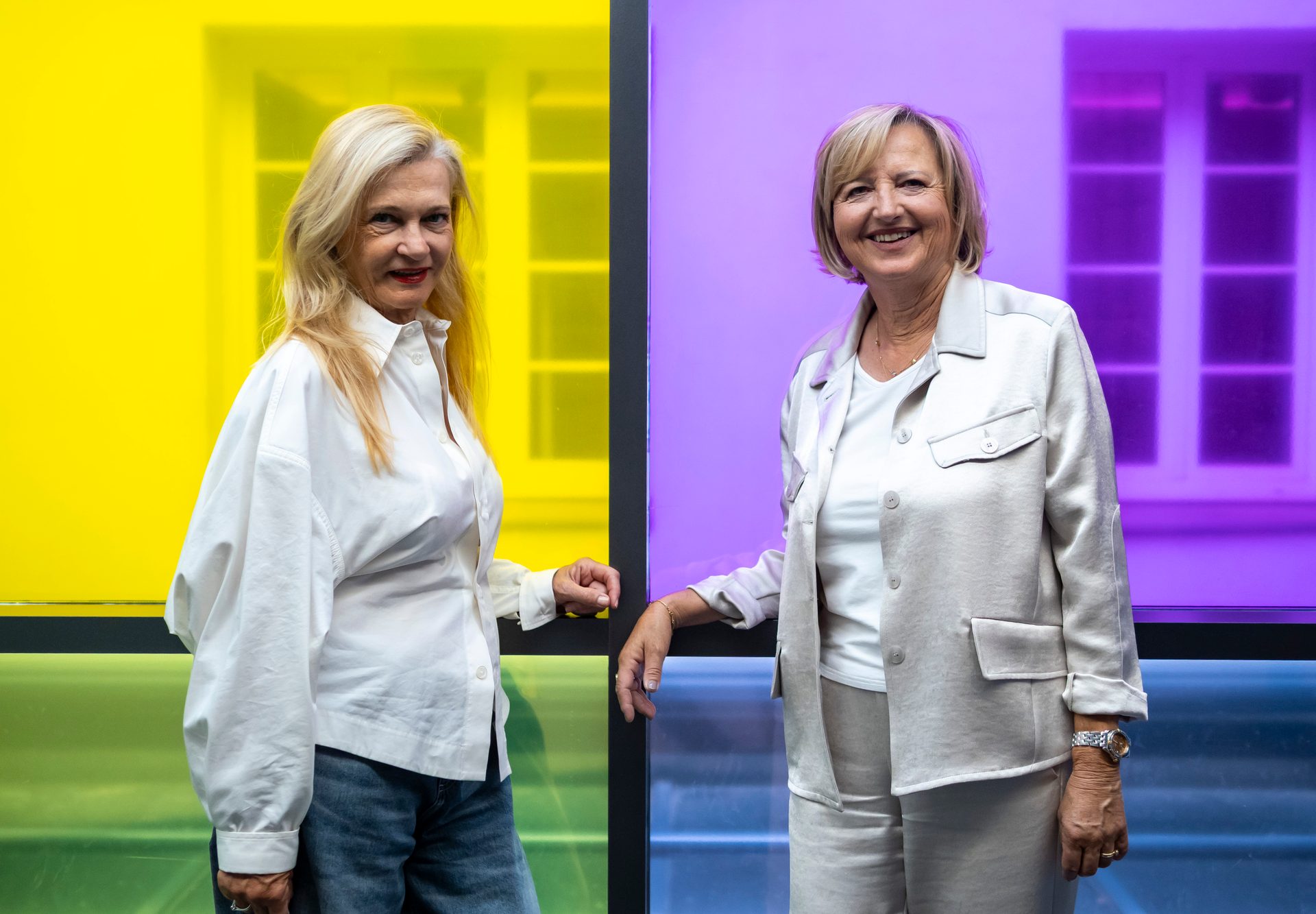 Two smiling women, one in a white shirt and the other in a light suit, stand against colorful panels.