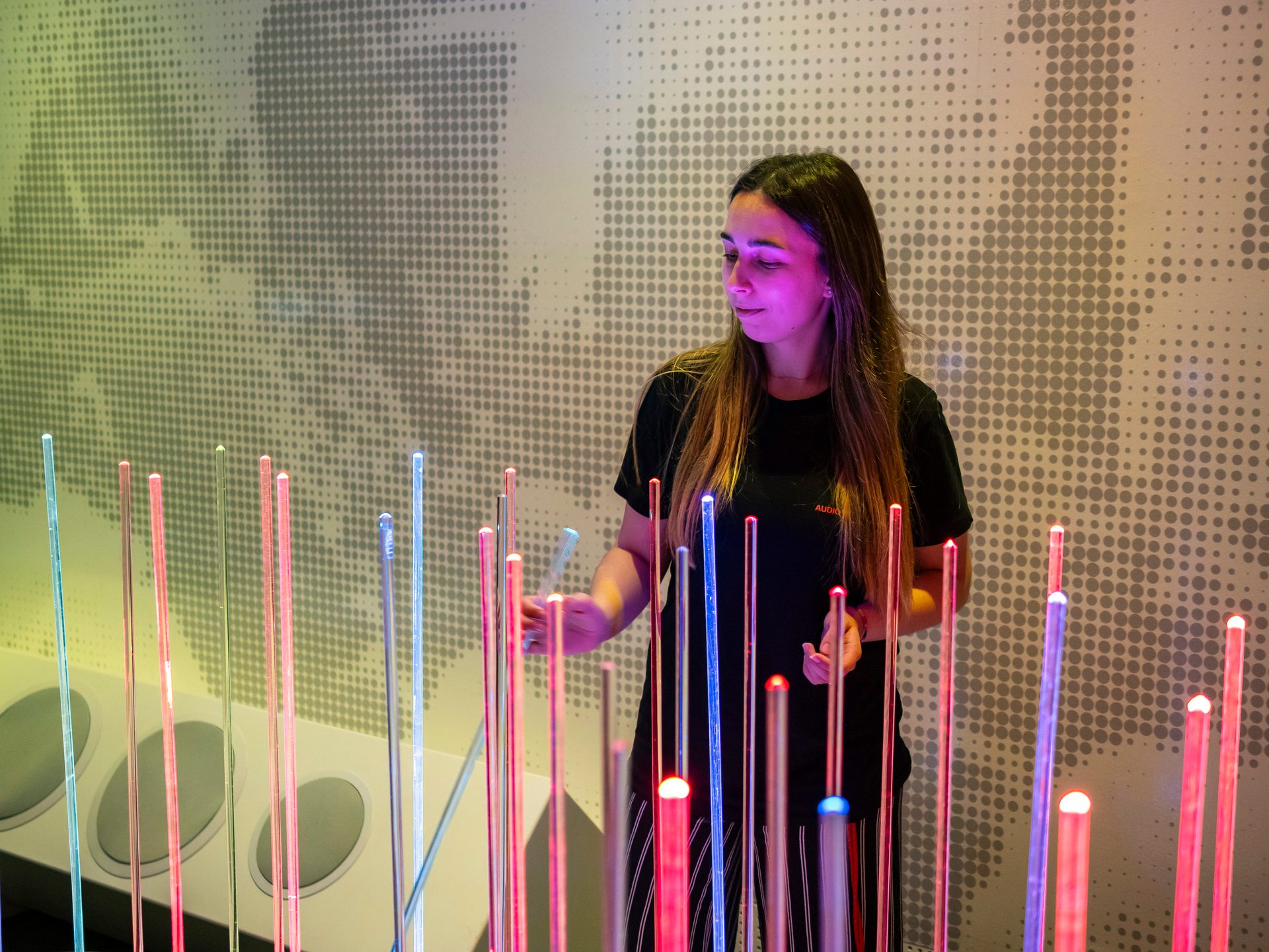 Woman interacts with glowing light rods in an art installation with a dotted wall.