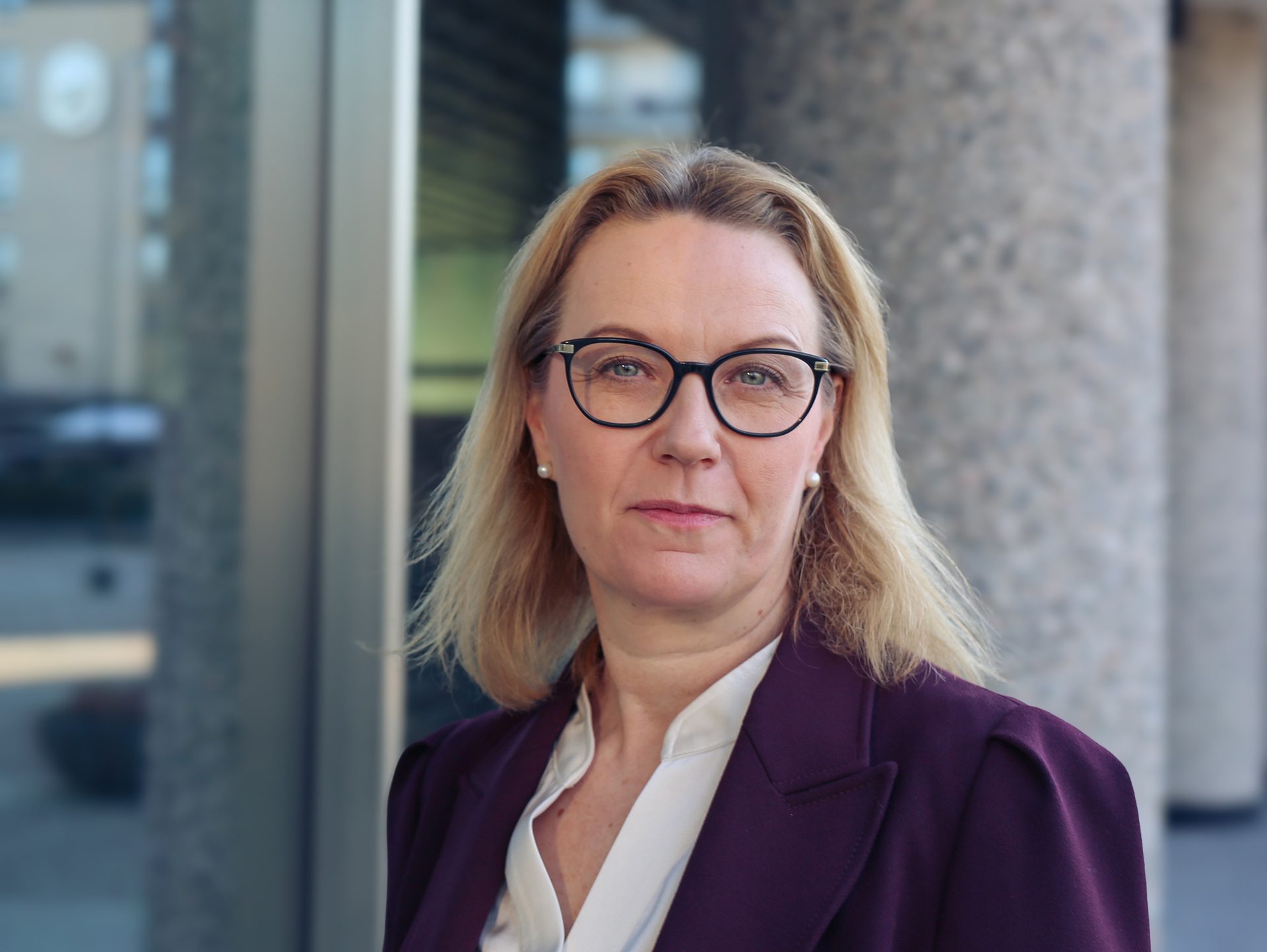 A confident woman with blonde hair and glasses, wearing a purple blazer, looking directly at the camera.