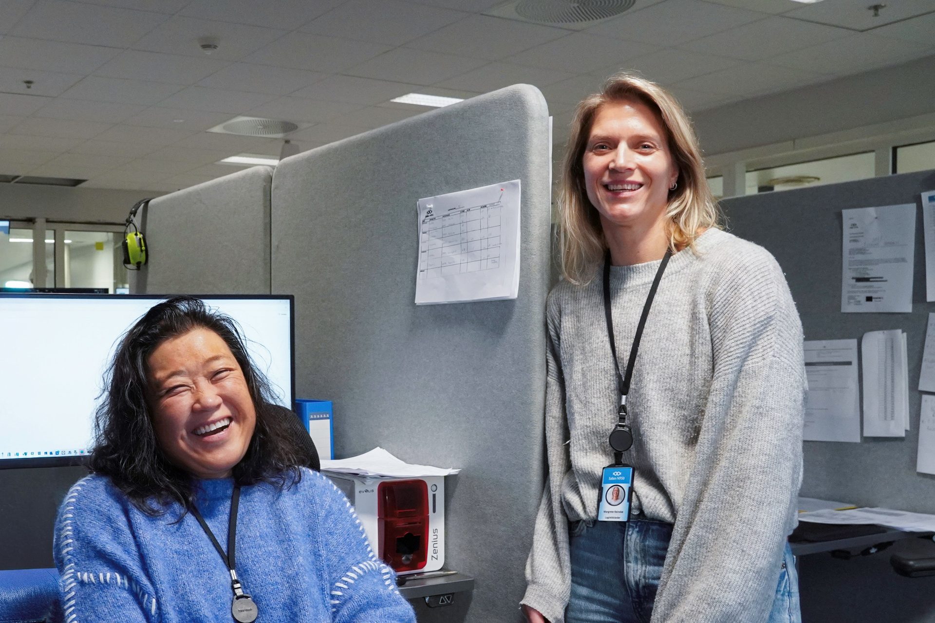 Two cheerful women, one laughing and one smiling, are pictured together in an office environment with cubicle walls.