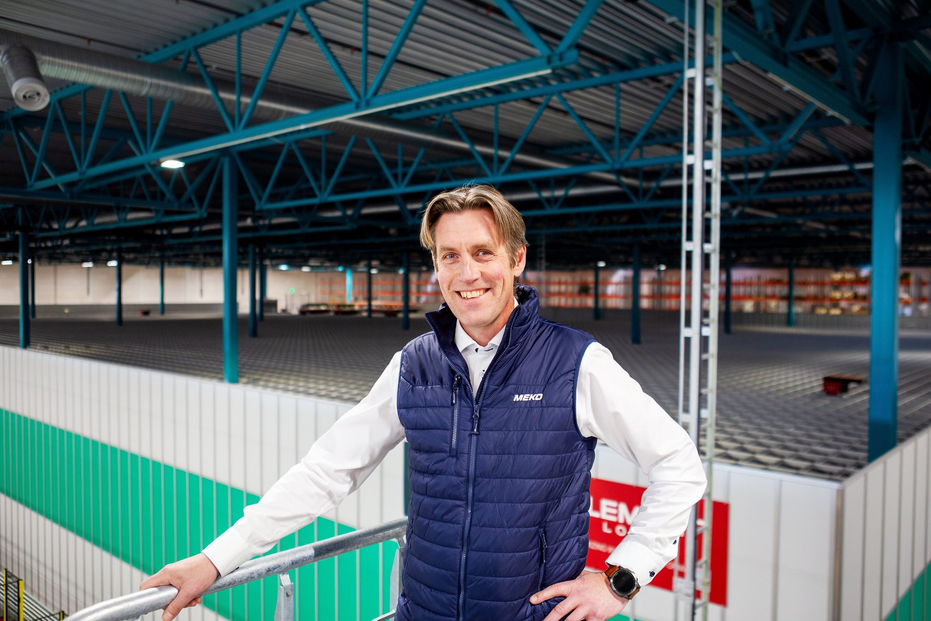 A smiling man in a blue vest stands in a large, empty logistics warehouse with blue support beams.