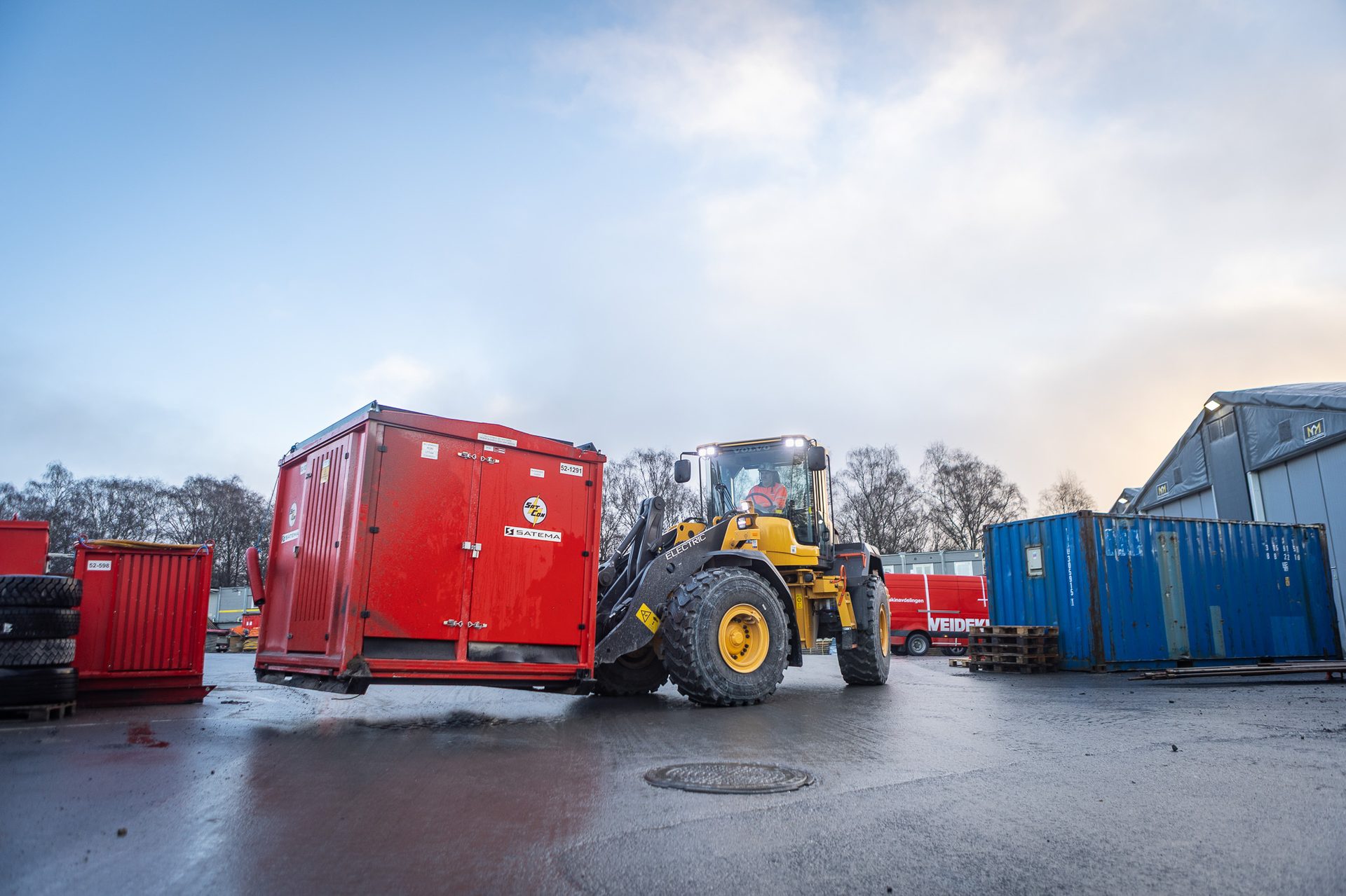 Yellow electric Volvo loader carrying a red container in an outdoor industrial area.