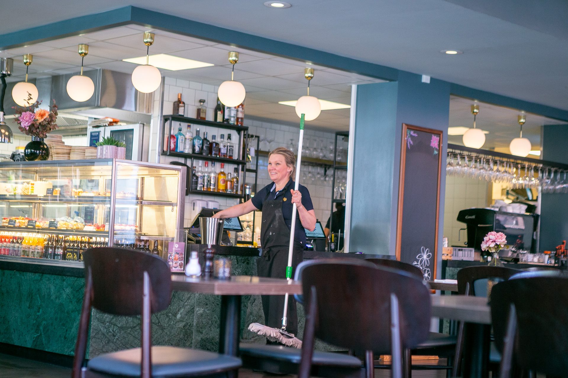 A smiling woman holds a mop in a cafe with tables and a counter.