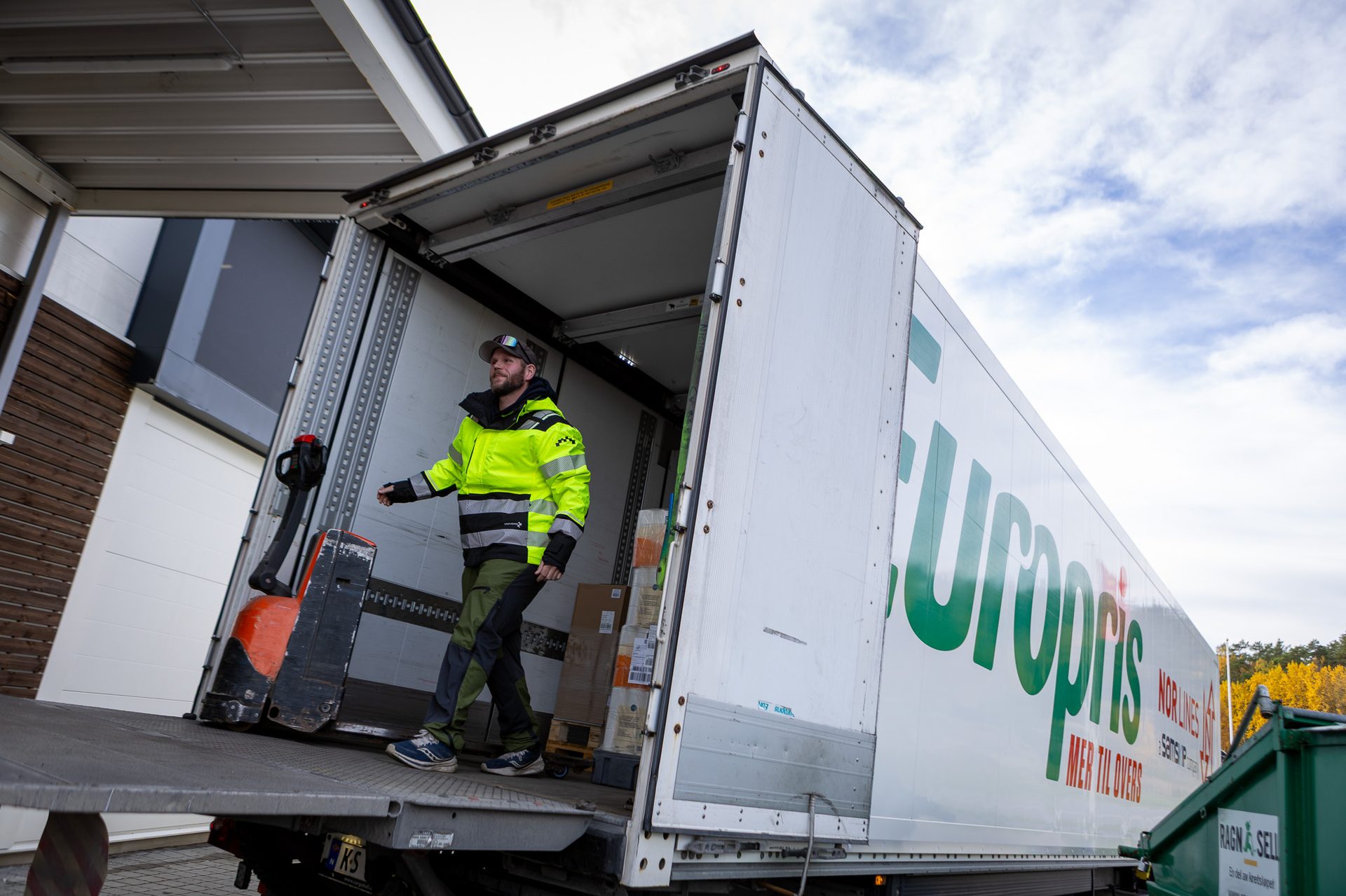 Man in neon jacket exits a Europris truck with a pallet jack, delivering goods.