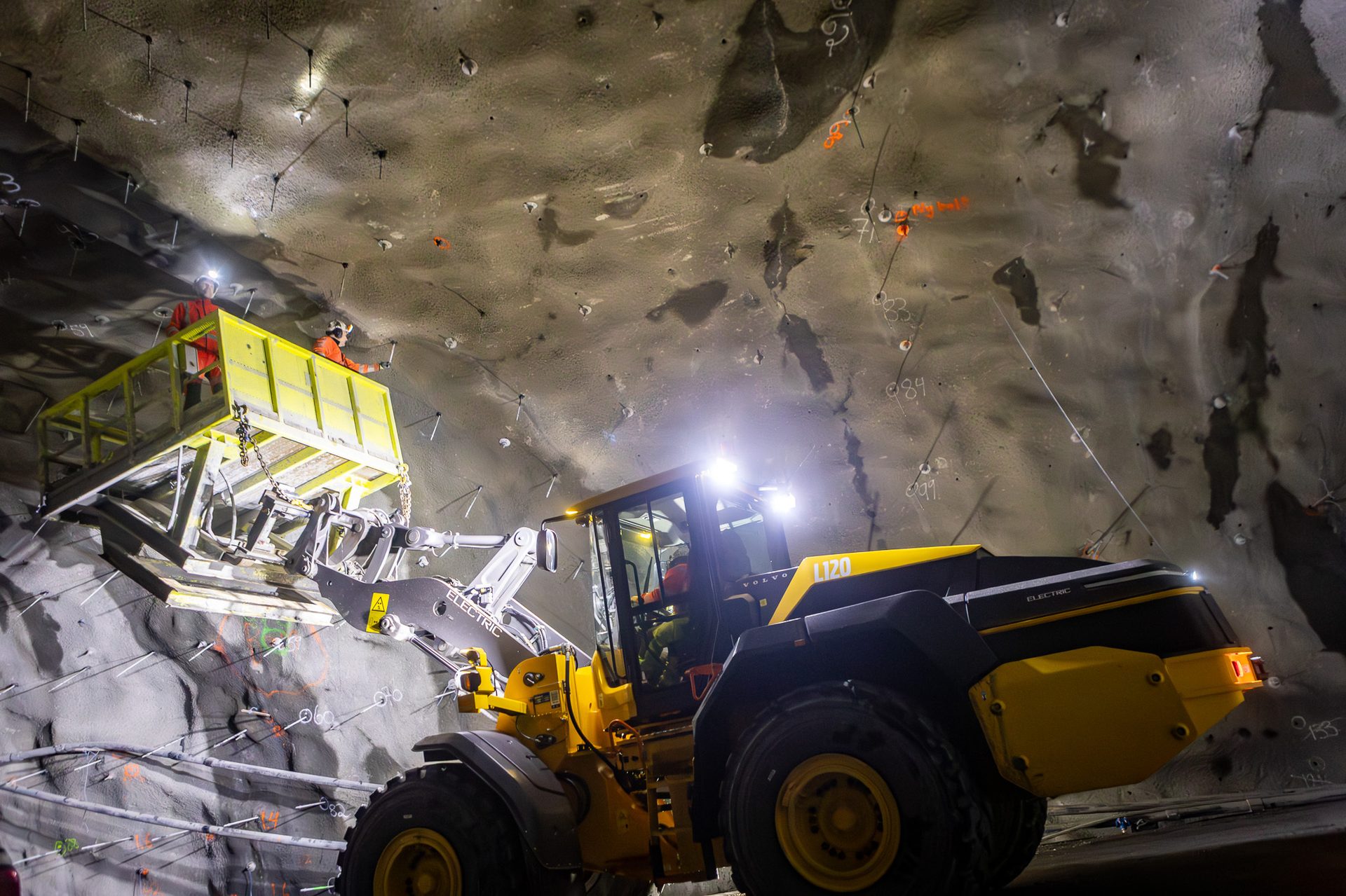 Electric Volvo loader with workers reinforcing an underground tunnel ceiling.