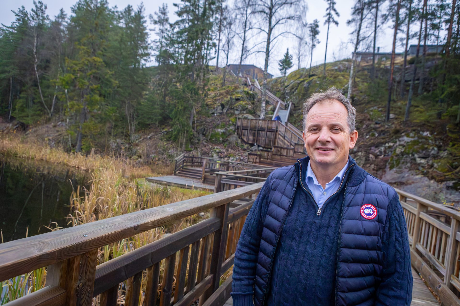 A smiling man in a blue vest stands on a wooden walkway, with a natural landscape and stairs behind him.