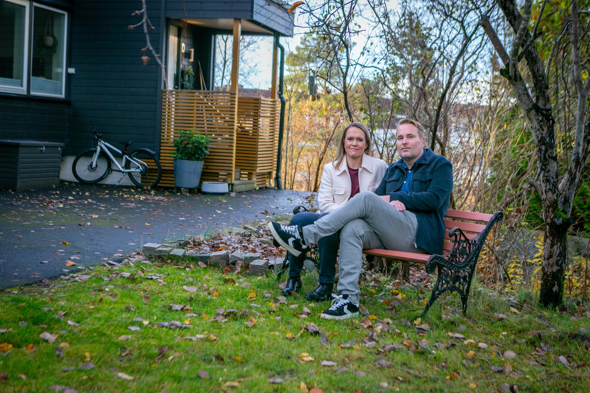 A man and woman sit on an outdoor bench in front of a house, surrounded by autumn trees.