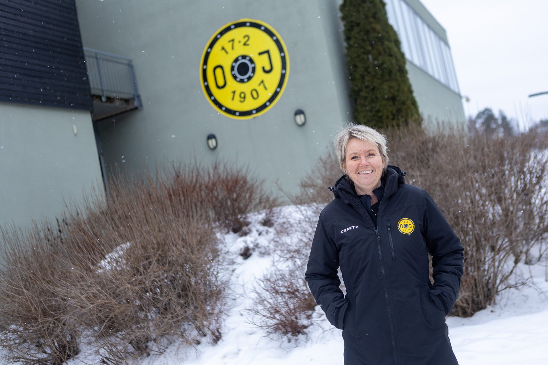 Smiling woman in a black jacket standing in snow, with a building and yellow logo behind her.