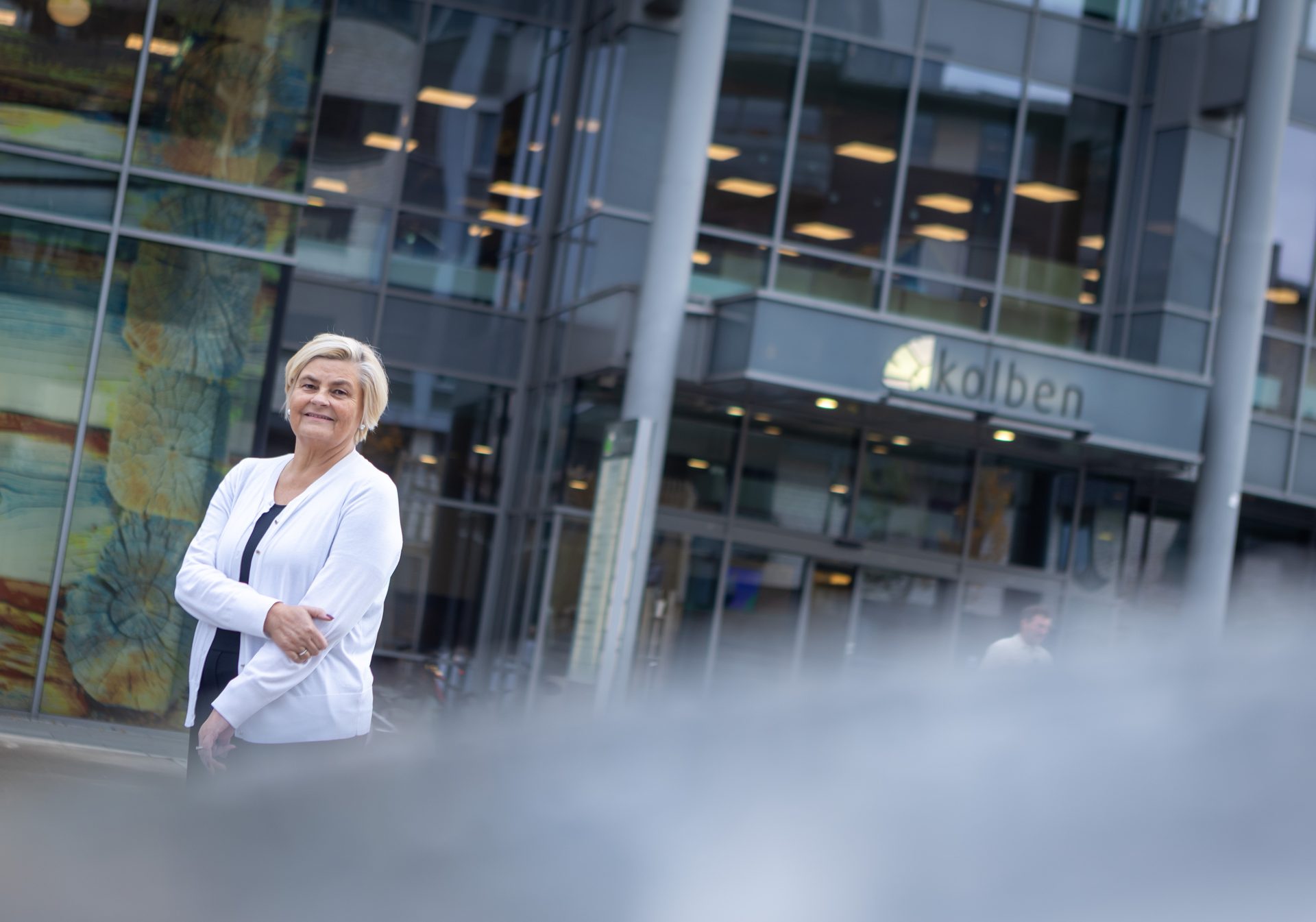 Smiling blonde woman in a white cardigan stands in front of a modern glass building with a "Kolben" sign.