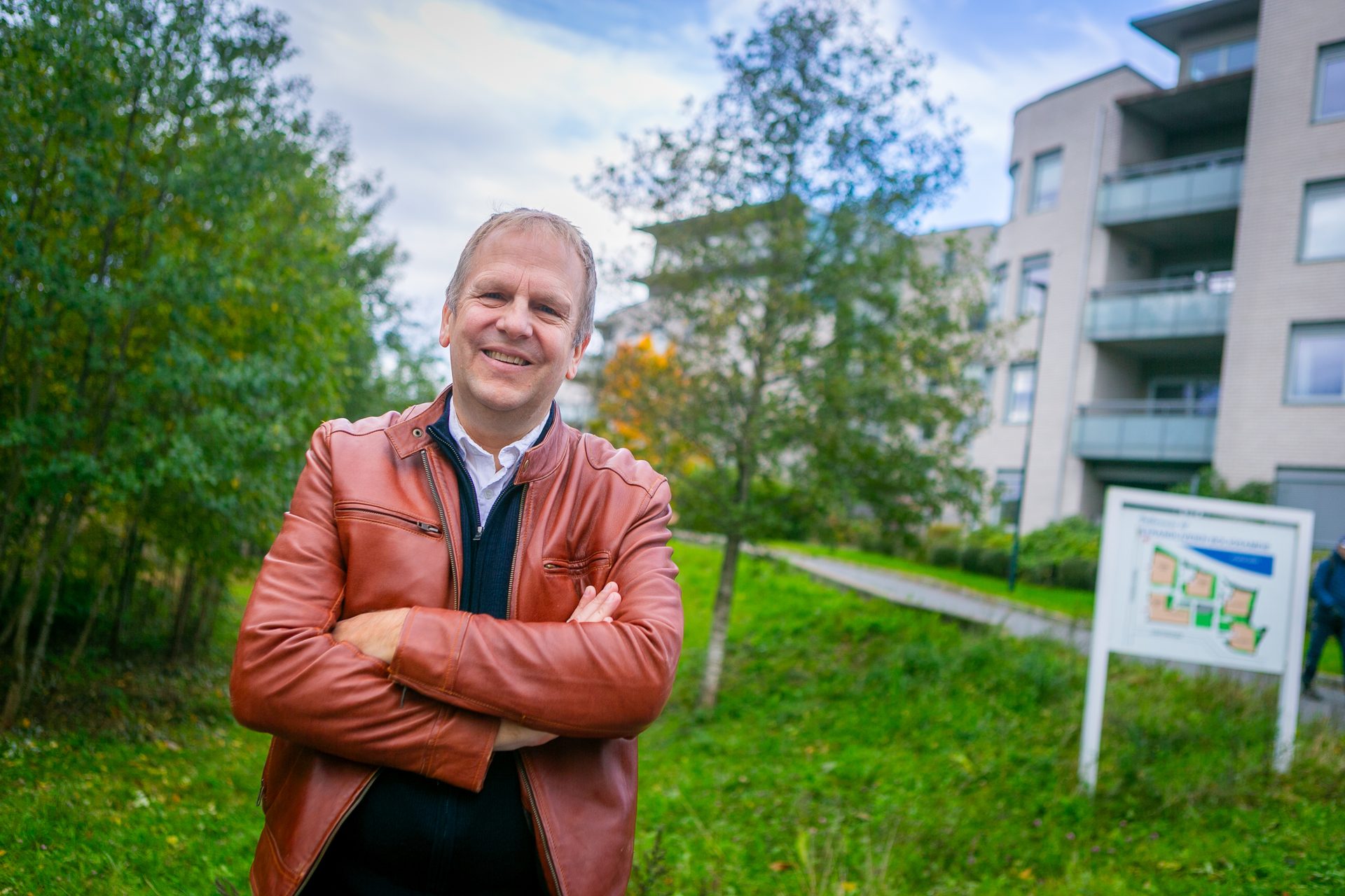 Smiling man in a brown leather jacket stands outdoors with trees and a building in the background.