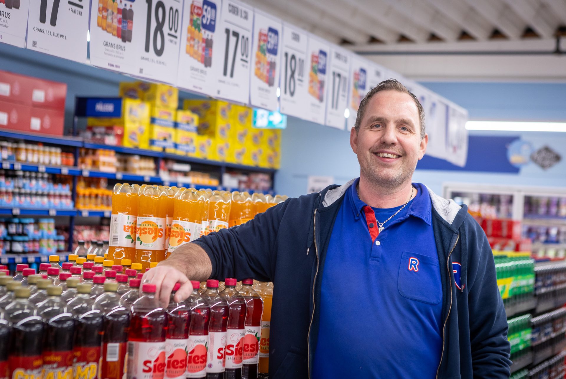 Smiling man in a blue shirt and hoodie in a supermarket beverage aisle, leaning on soda bottles.