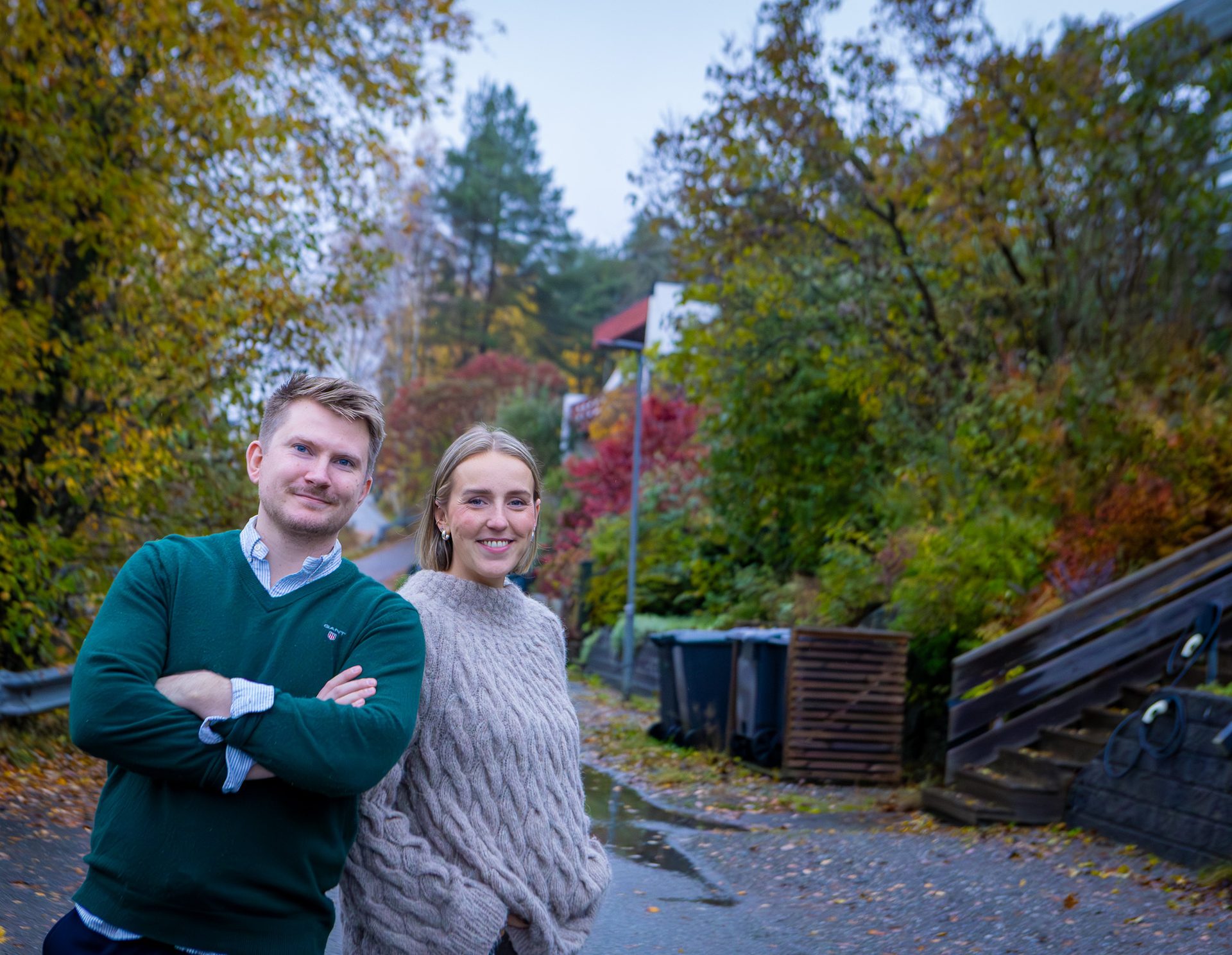 A smiling man and woman stand on an autumn street with colorful trees and houses in the background.