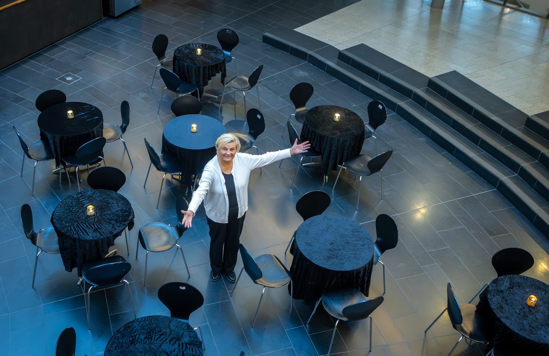 Smiling woman with arms wide among black draped tables with candles and chairs in a modern venue.