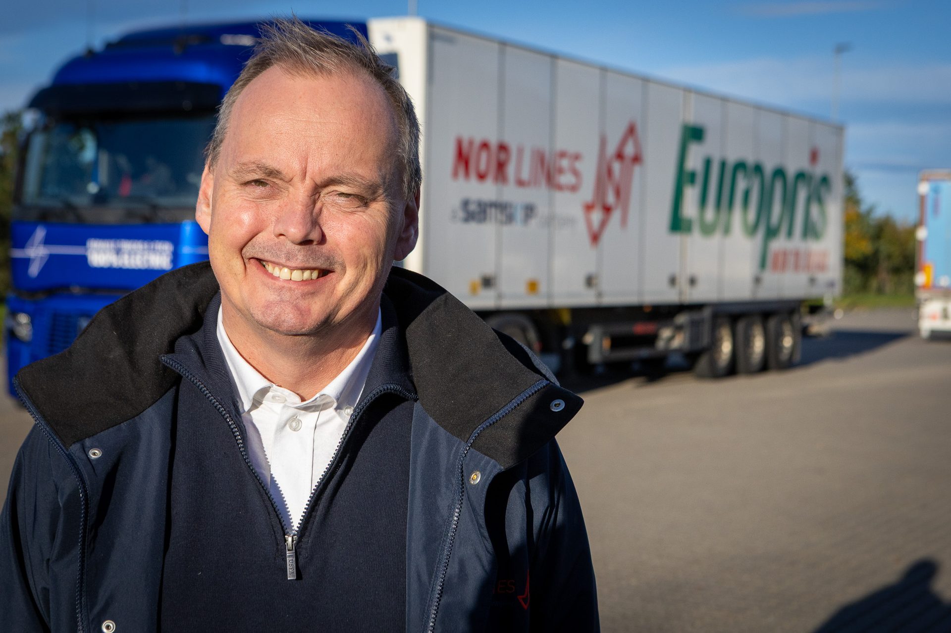 Smiling man in blue jacket with trucks featuring "NOR LINES" and "Europris" logos in background.