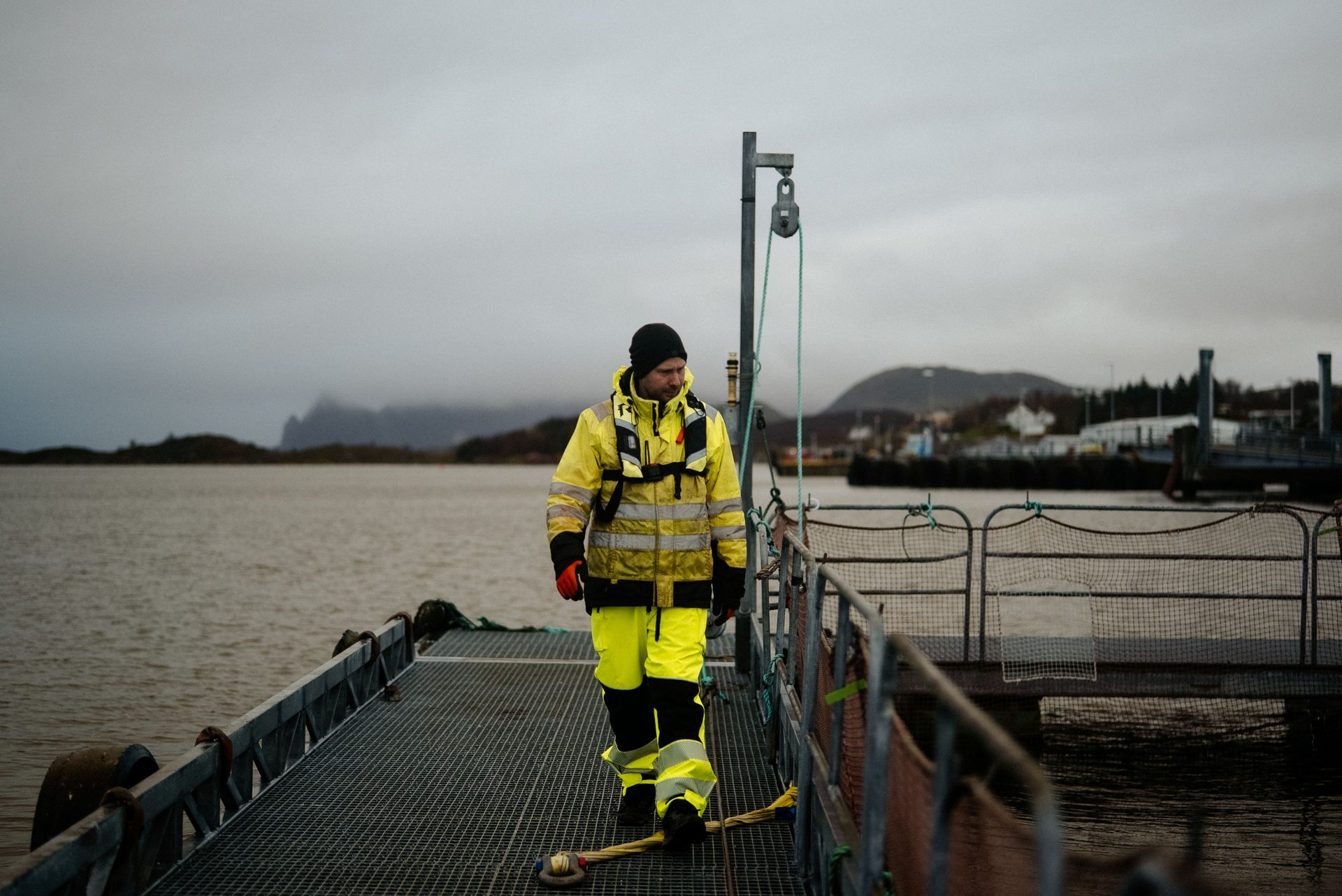 Man in bright yellow safety gear walks on a metal pier with water and mountains under a cloudy sky.