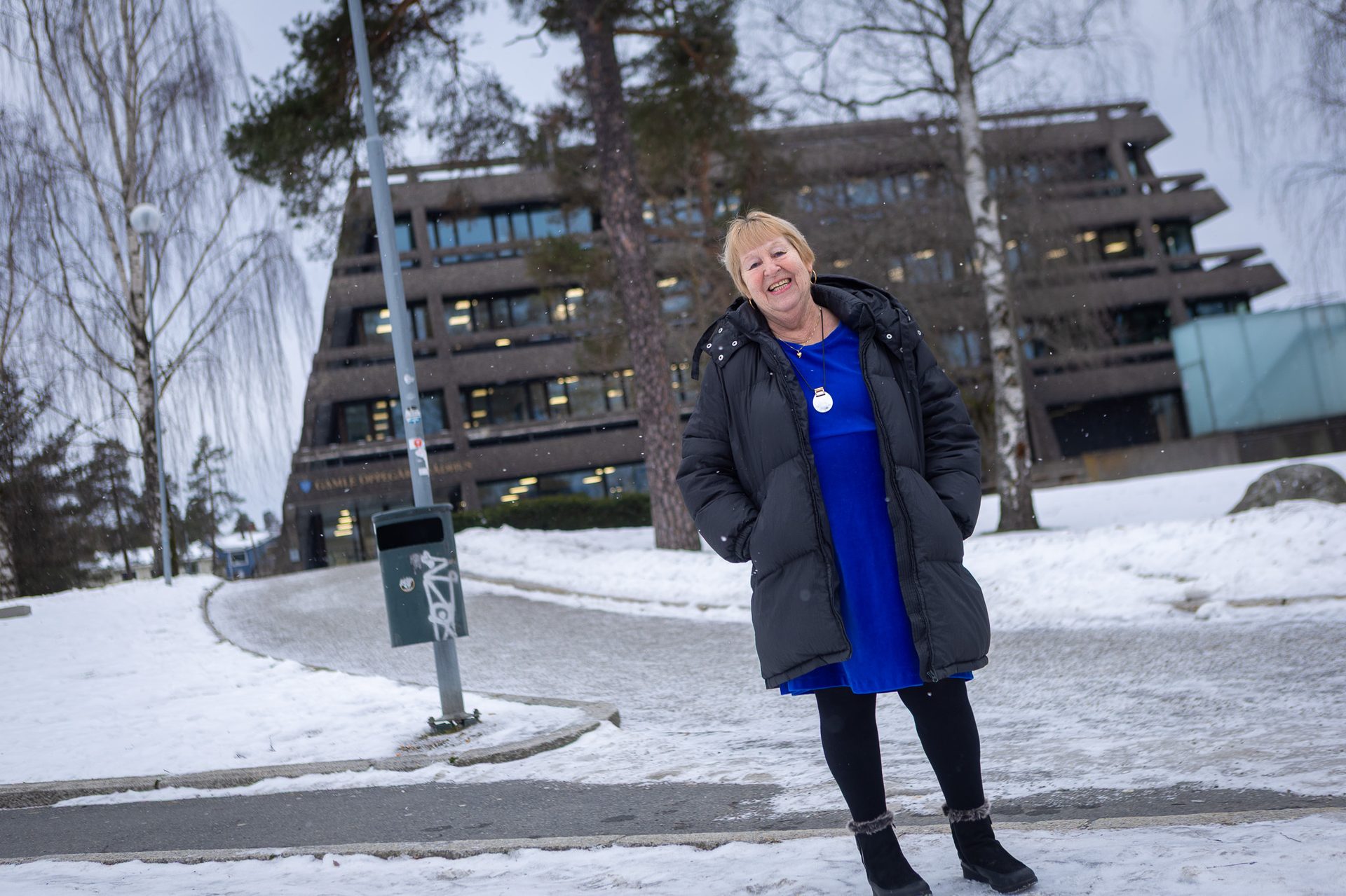 Smiling woman in black puffer coat and blue dress standing in snow.