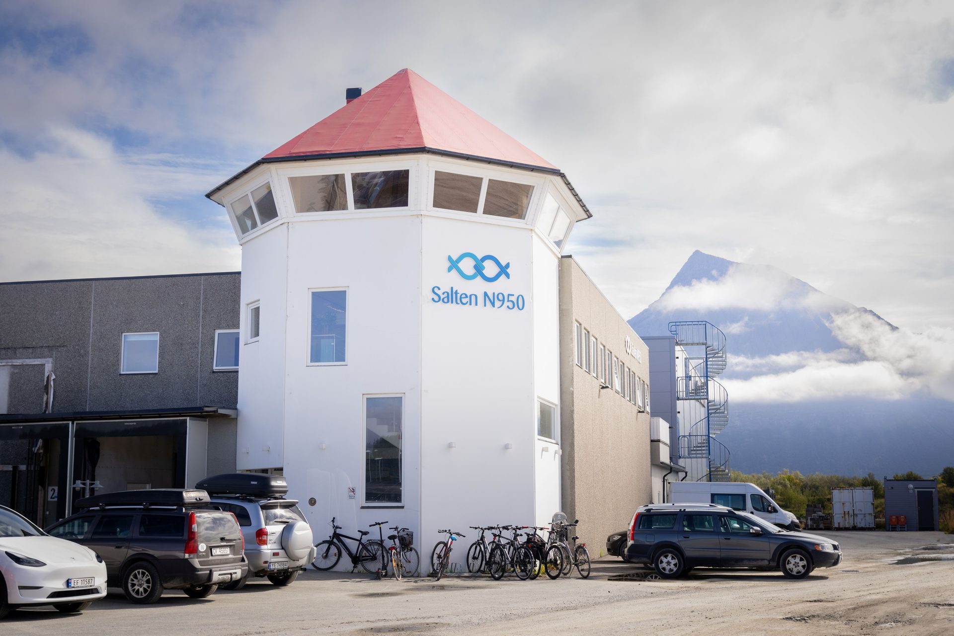 White building with a red roof and "Salten N950" logo, parked cars, bicycles, and a cloud-covered mountain.