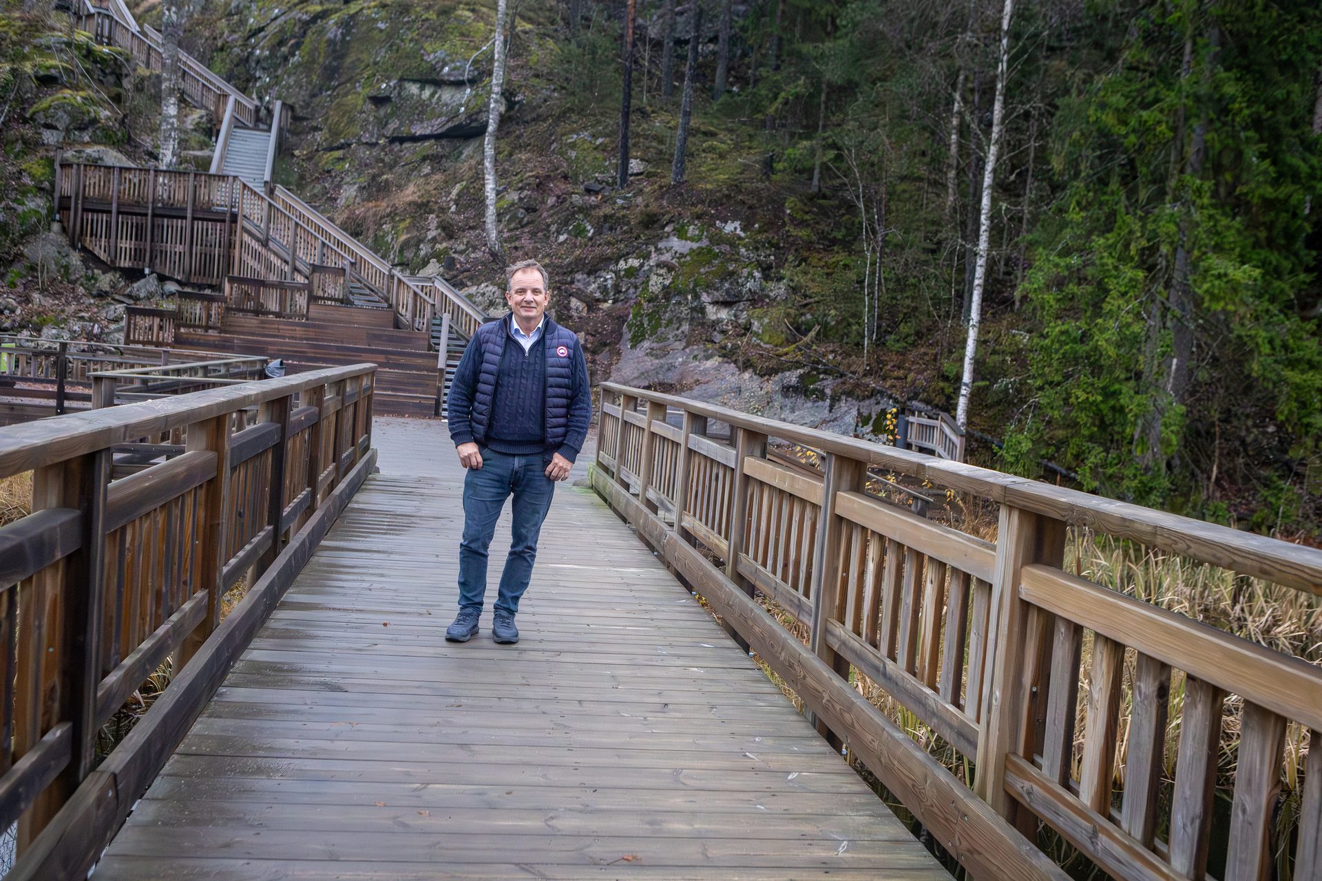 Man on a wooden boardwalk, with wooden stairs leading up a steep, forested hillside.