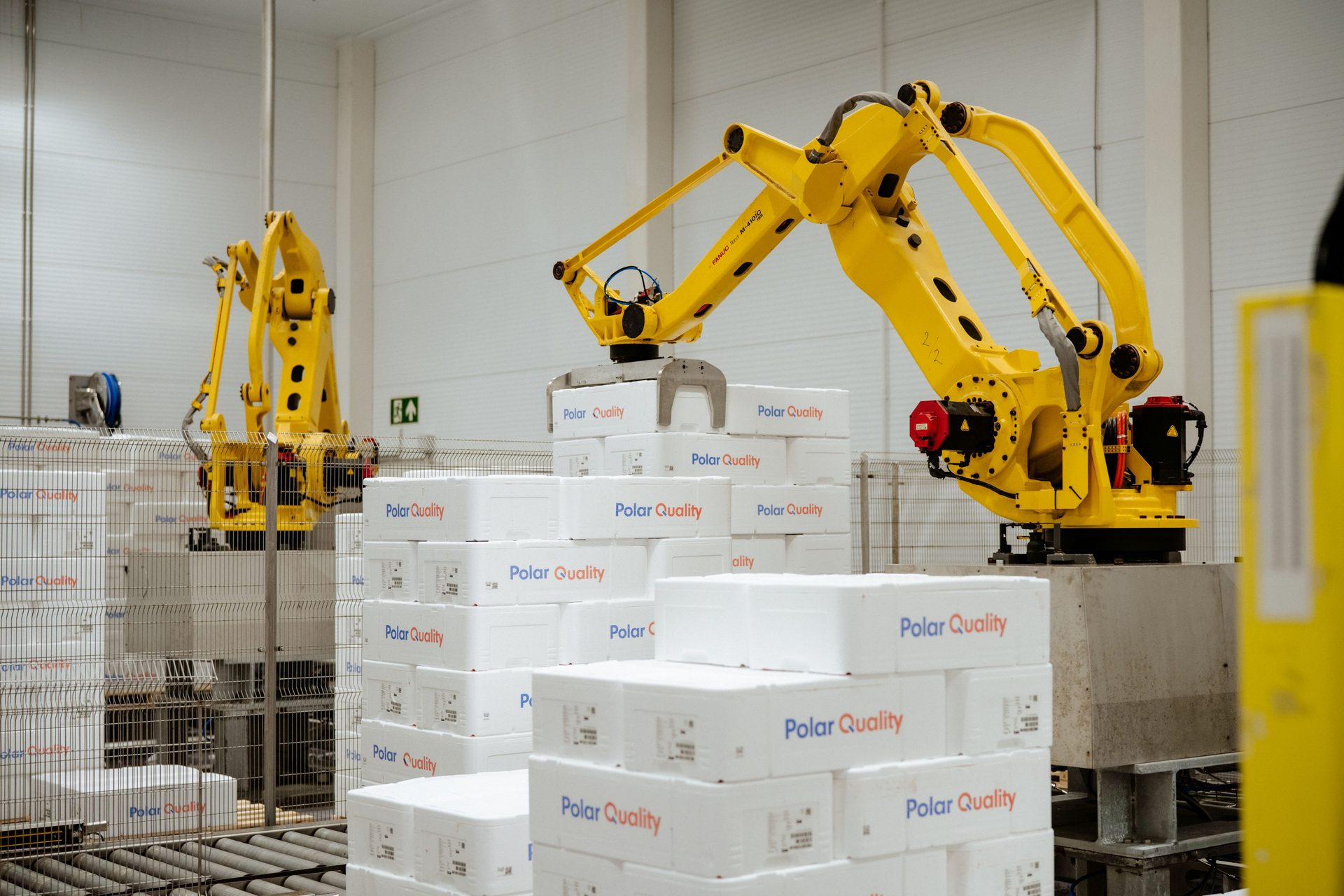 Two yellow robotic arms stacking white "Polar Quality" boxes in an automated warehouse.