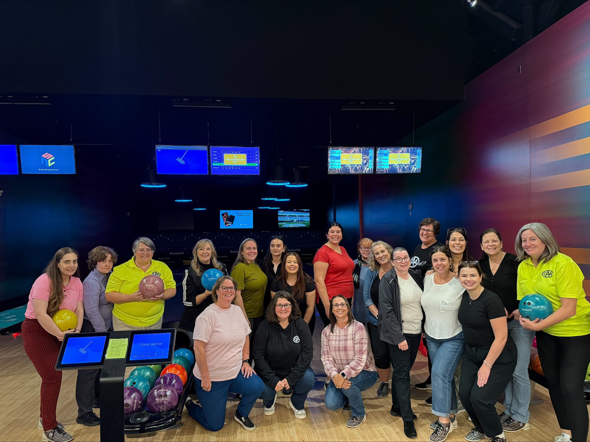 A diverse group of women pose with bowling balls in a modern bowling alley, smiling at the camera.