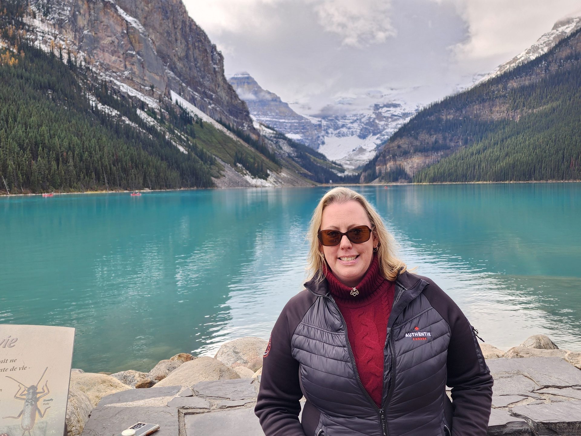 Smiling woman at scenic Lake Louise with turquoise water, snowy mountains, and trees.