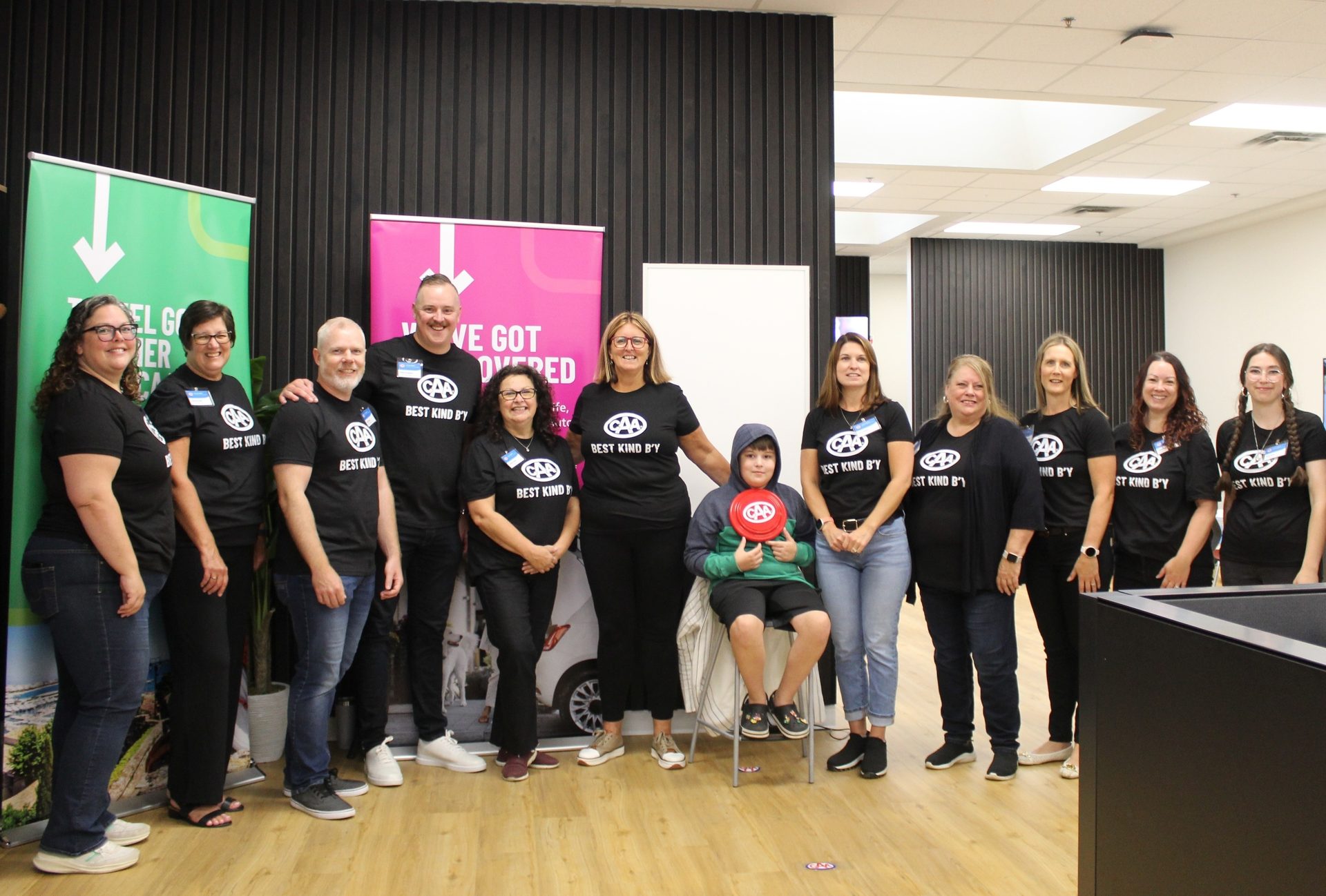 A group of people and a child in black "CAA BEST KIND B'Y" t-shirts stand together in a room.