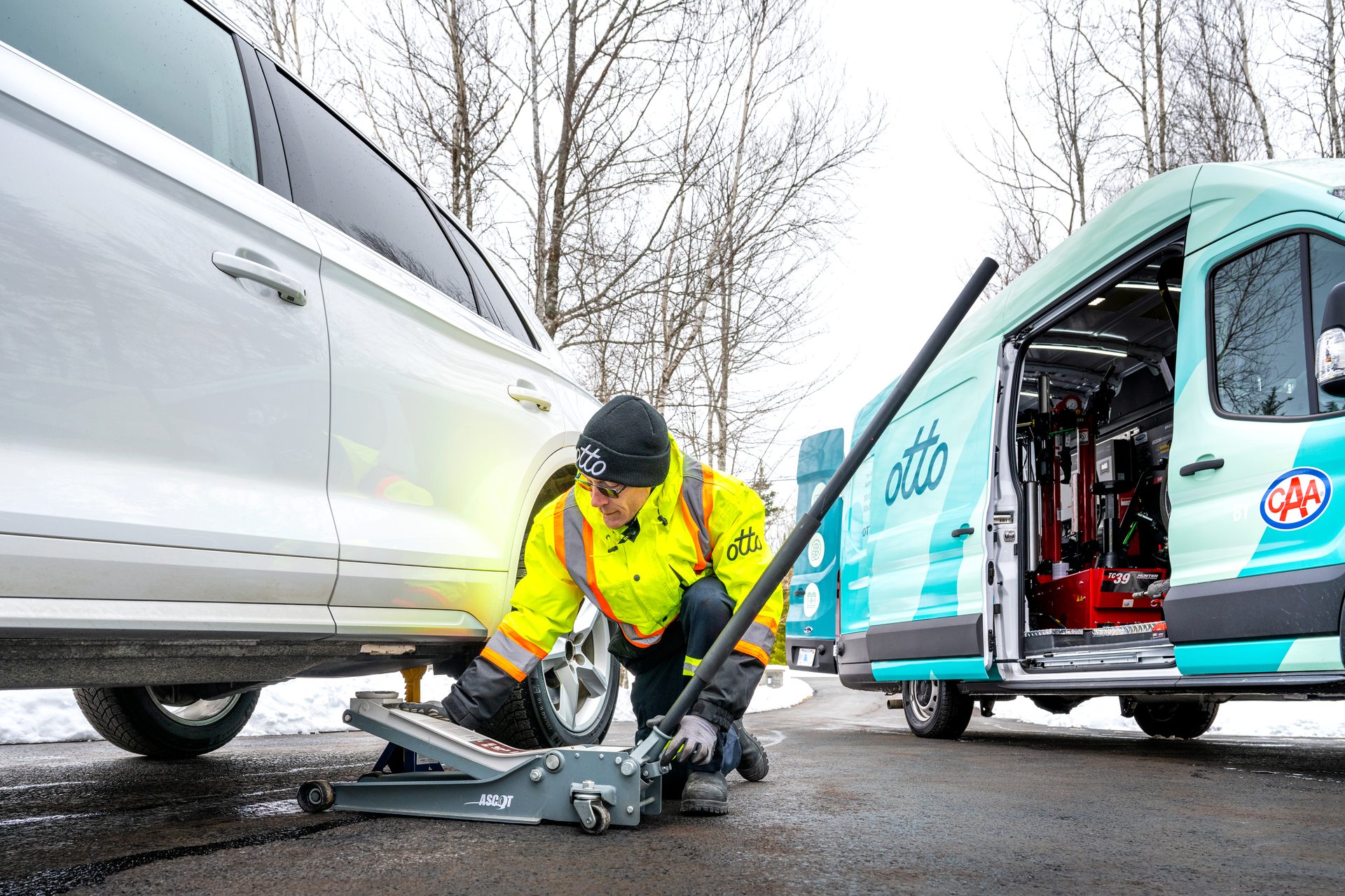 Man in hi-vis jacket jacks up white car; "Otto" & "CAA" service van nearby.