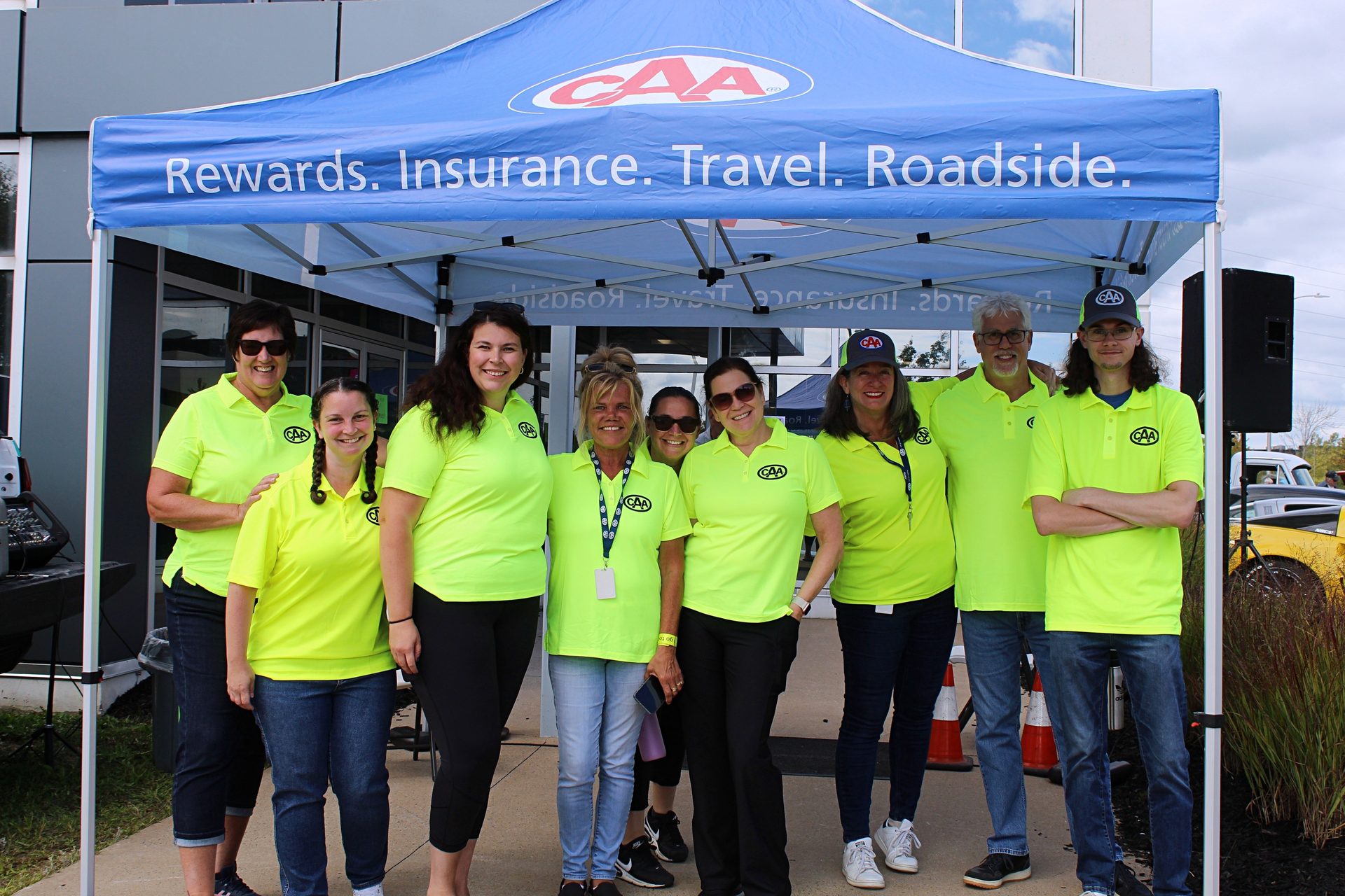 Nine smiling people in neon yellow CAA shirts under a blue tent.