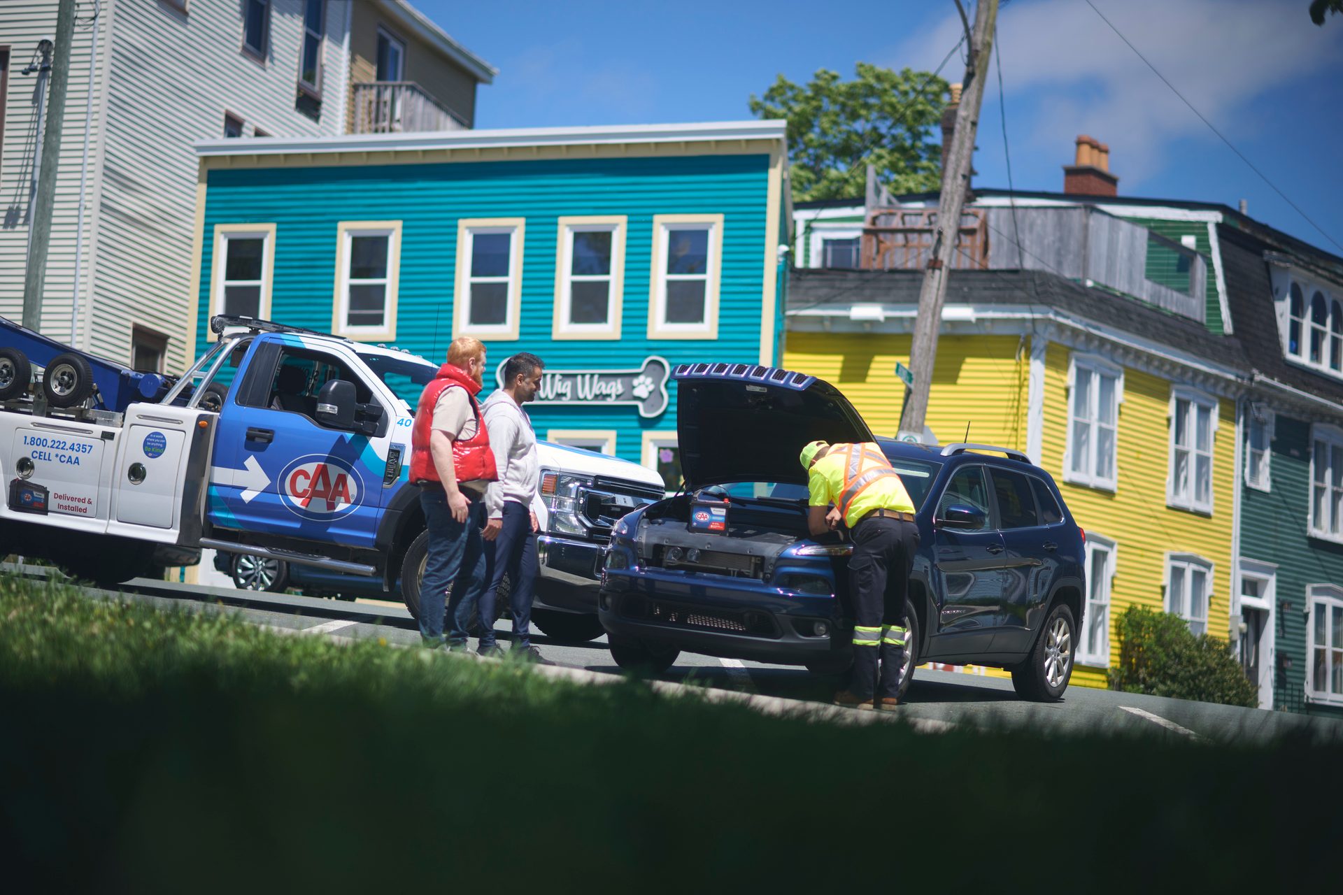 CAA tow truck and technician inspecting a car engine on a street with colorful houses.