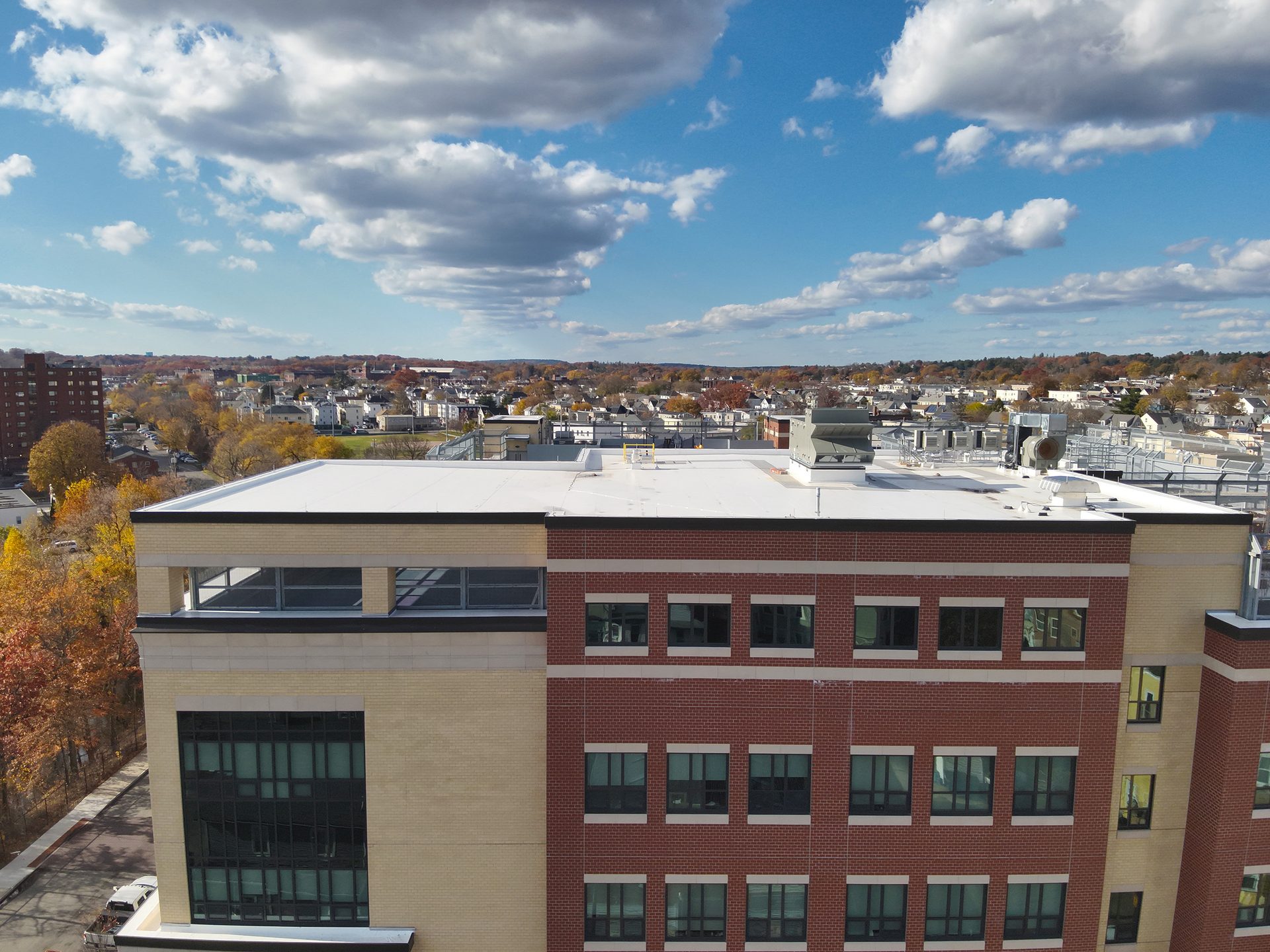 Aerial view of a large building with red brick and beige sections, overlooking a town and autumn trees under a cloudy sky.