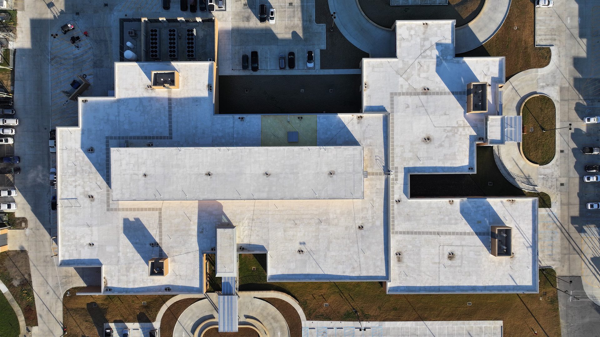 Aerial view of a sprawling modern building with light flat roofs, surrounded by parking and landscaping.