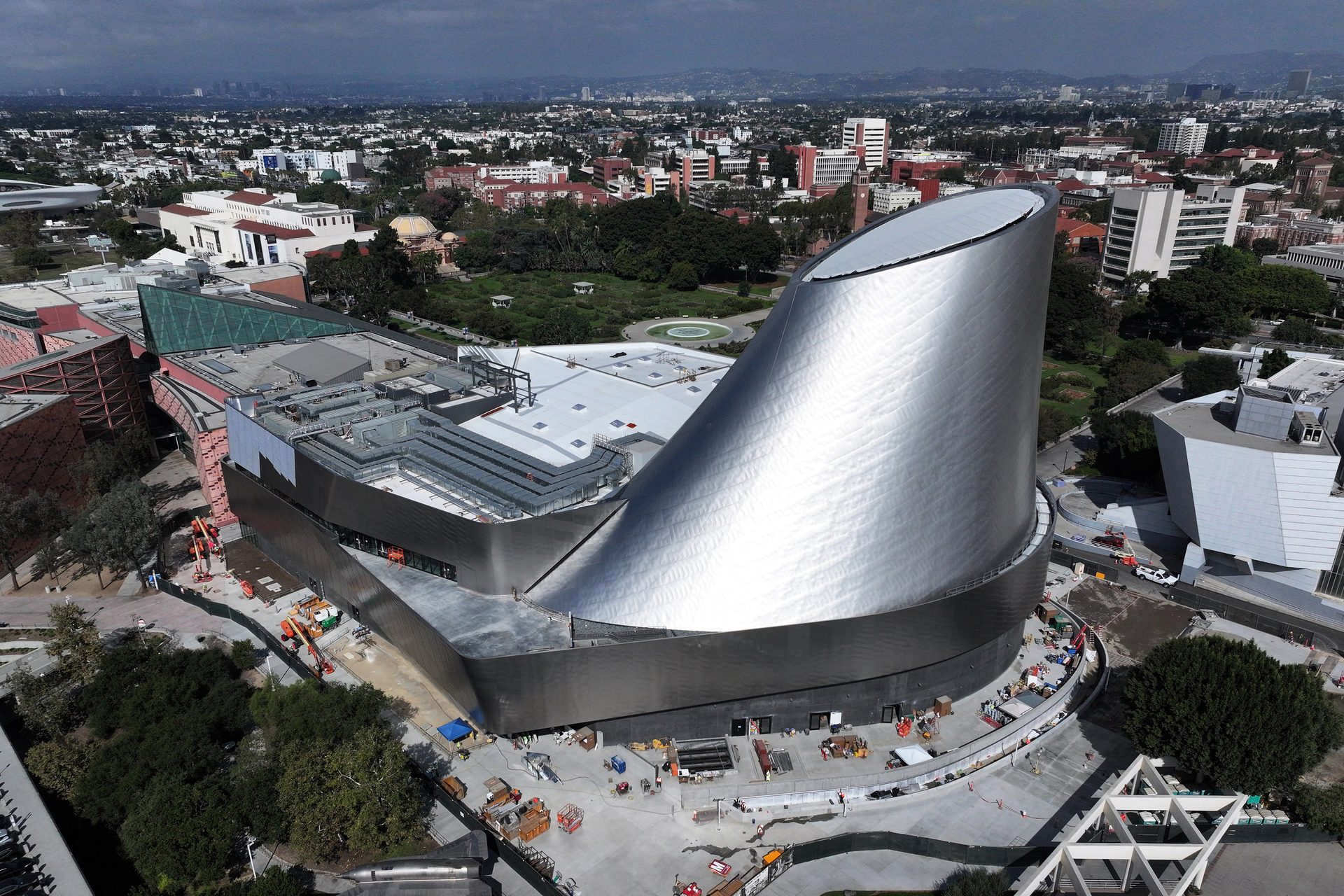 Aerial view of a striking silver-clad museum building under construction, surrounded by a cityscape.