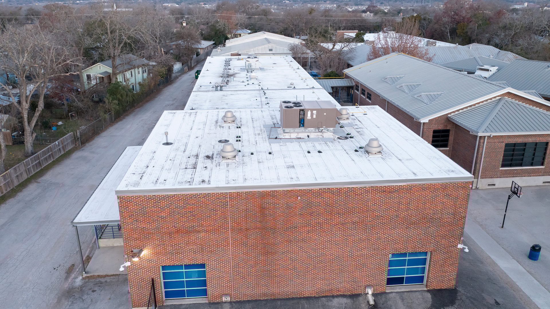 Aerial view of brick school building with white flat roof, HVAC, adjacent metal-roofed buildings, and an alley.