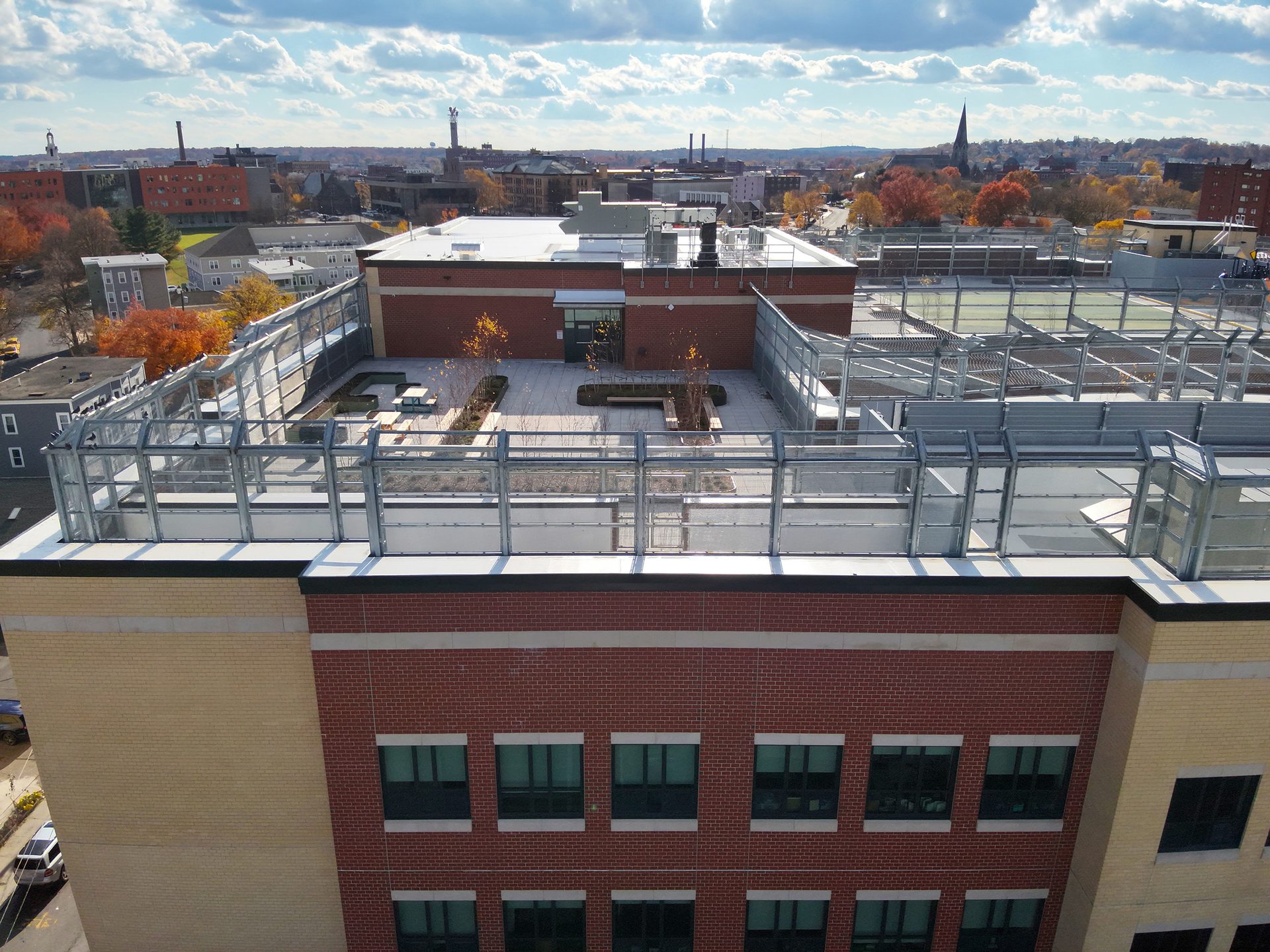 Rooftop garden with glass barriers on a brick building, city skyline with autumn trees in background.