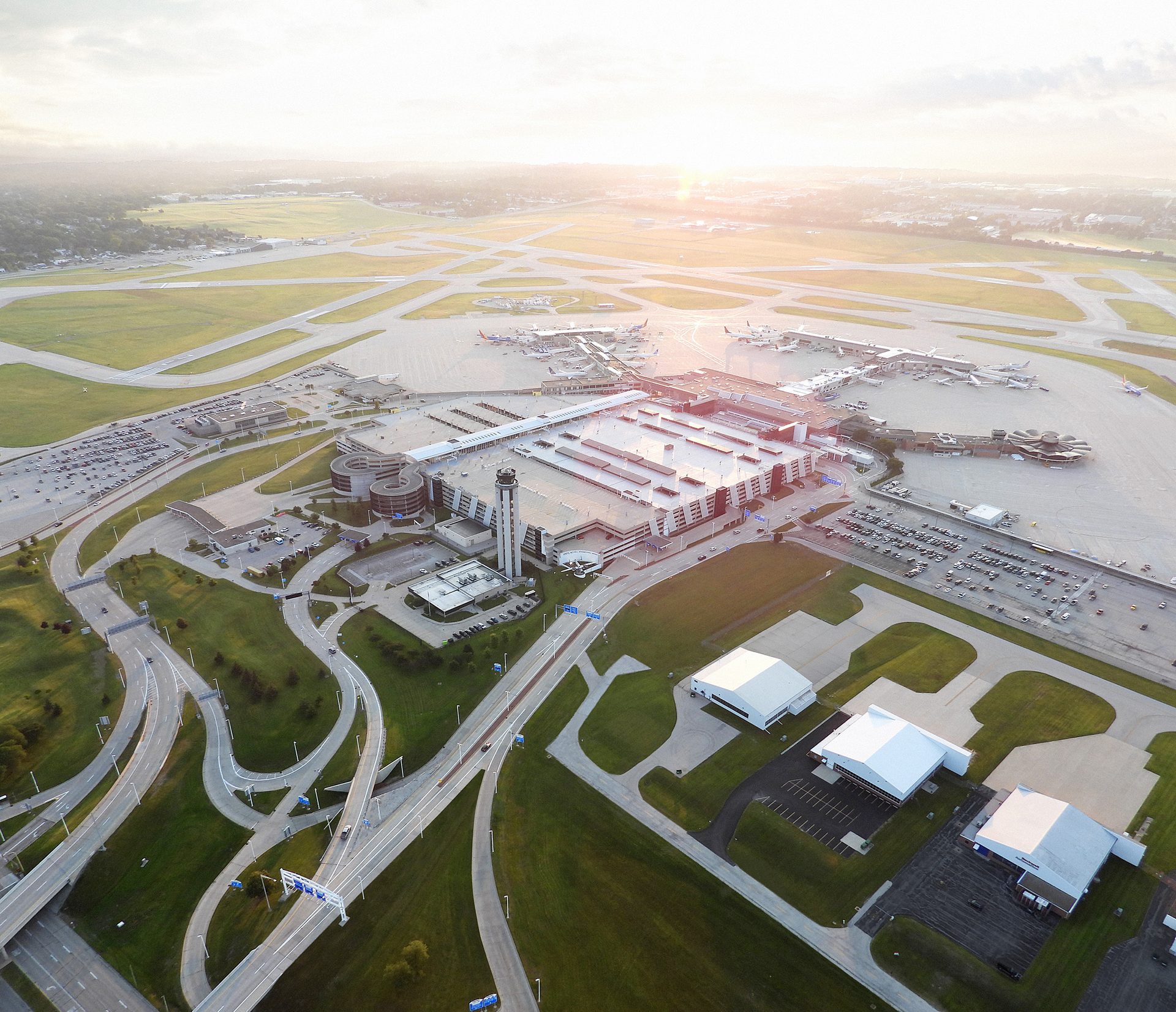 Aerial view of a large airport at sunset, showing the terminal, runways, parked aircraft, and surrounding roads.