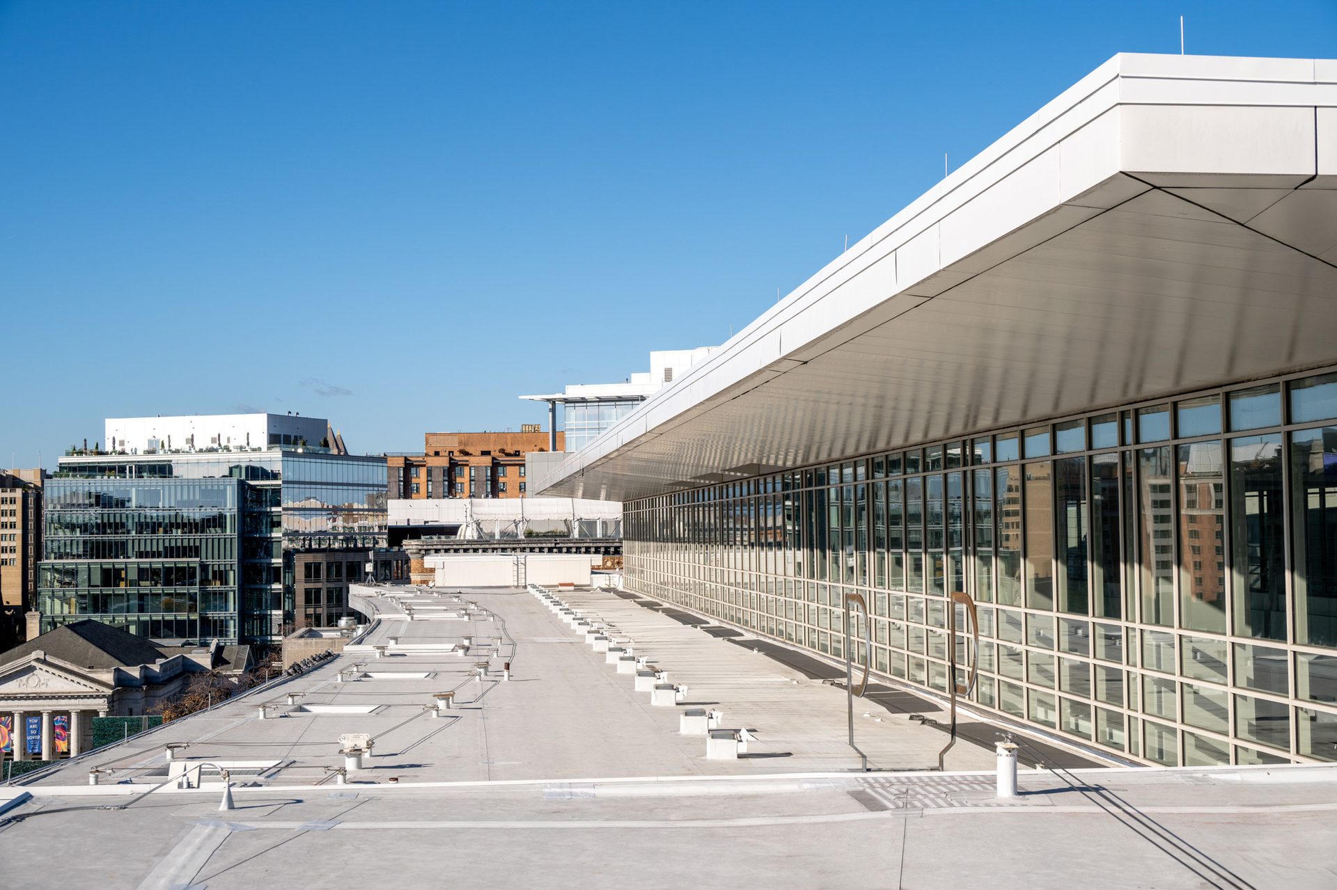 Modern building with glass facade and white overhang, seen from a rooftop. Cityscape and blue sky.