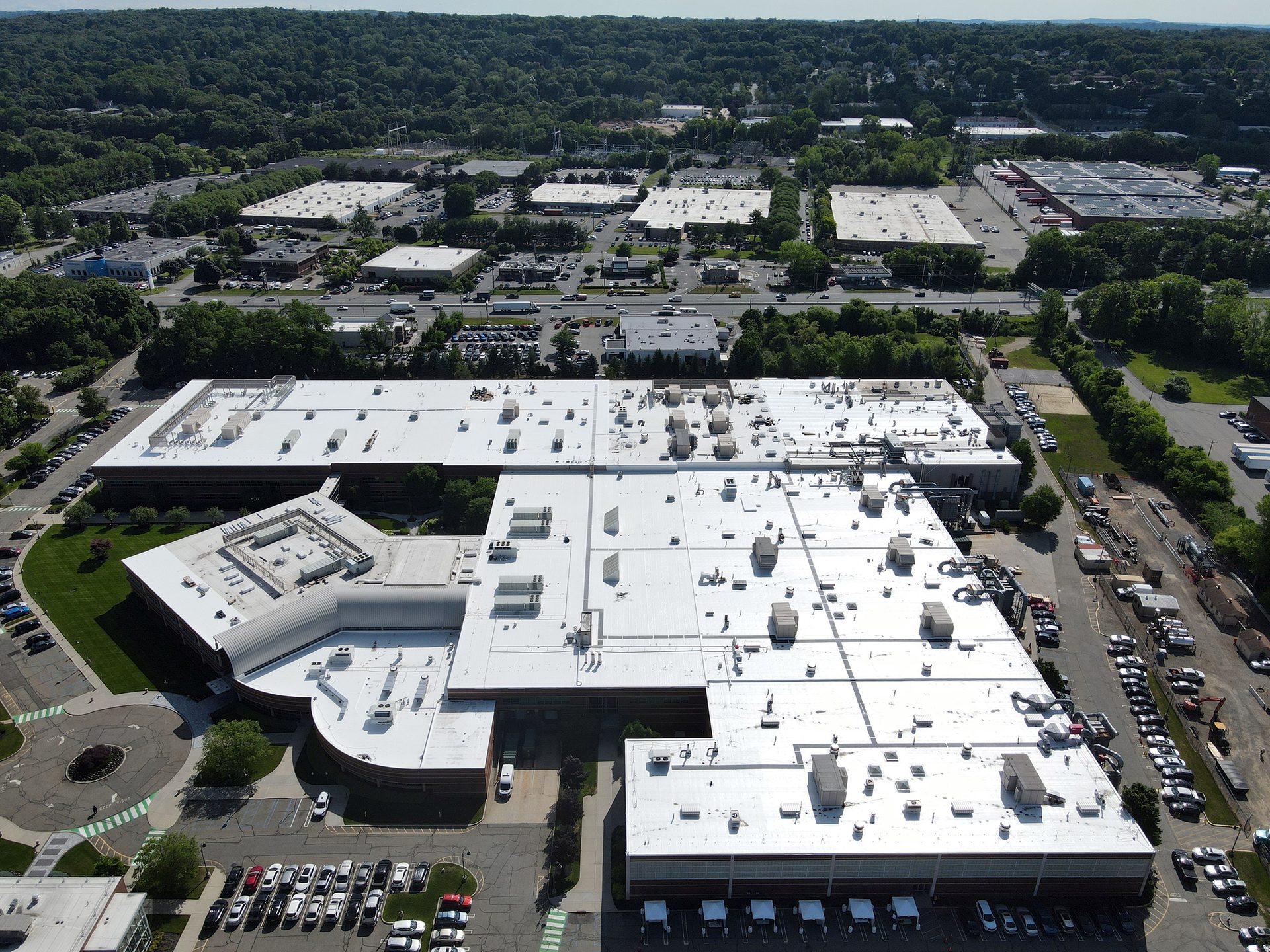 Aerial view of a large industrial complex with white roofs, surrounded by parking lots, roads, and green forests.