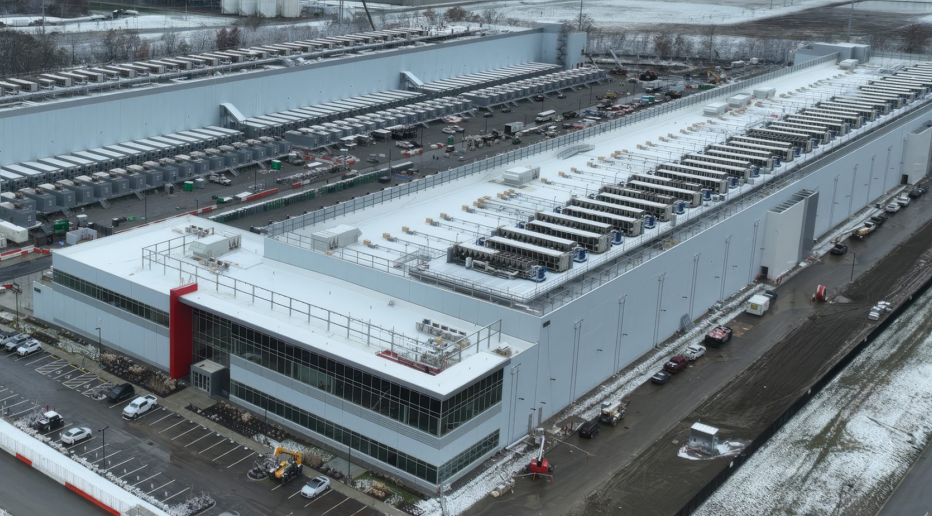 Aerial view of a large modern industrial or data center complex with multiple buildings, rooftop units, and loading docks, partially covered in snow.