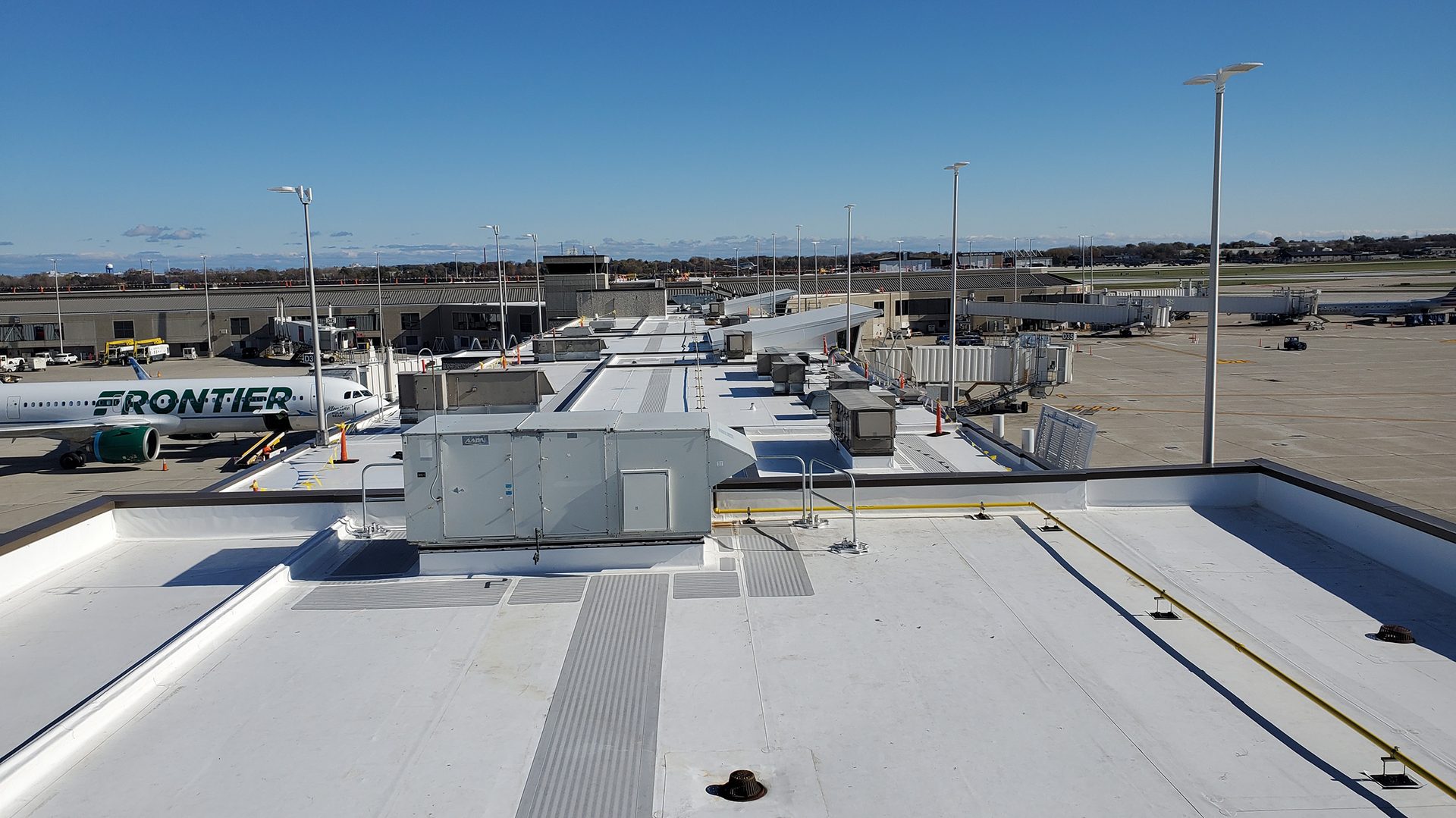 Aerial view of an airport tarmac with a Frontier plane, jet bridges, and terminal, over a rooftop with HVAC units.