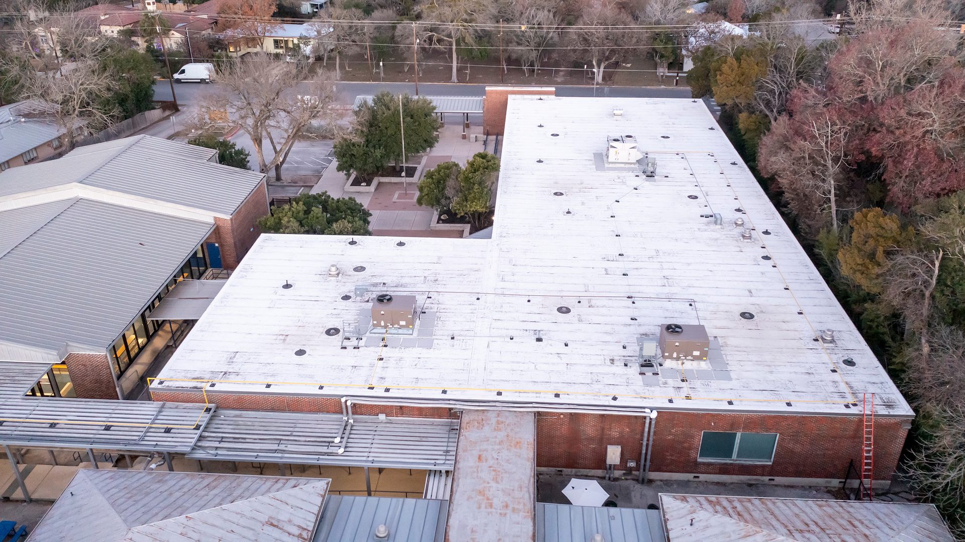 Aerial view of a school or commercial building with large white flat roofs and grey metal roofs, surrounded by trees.