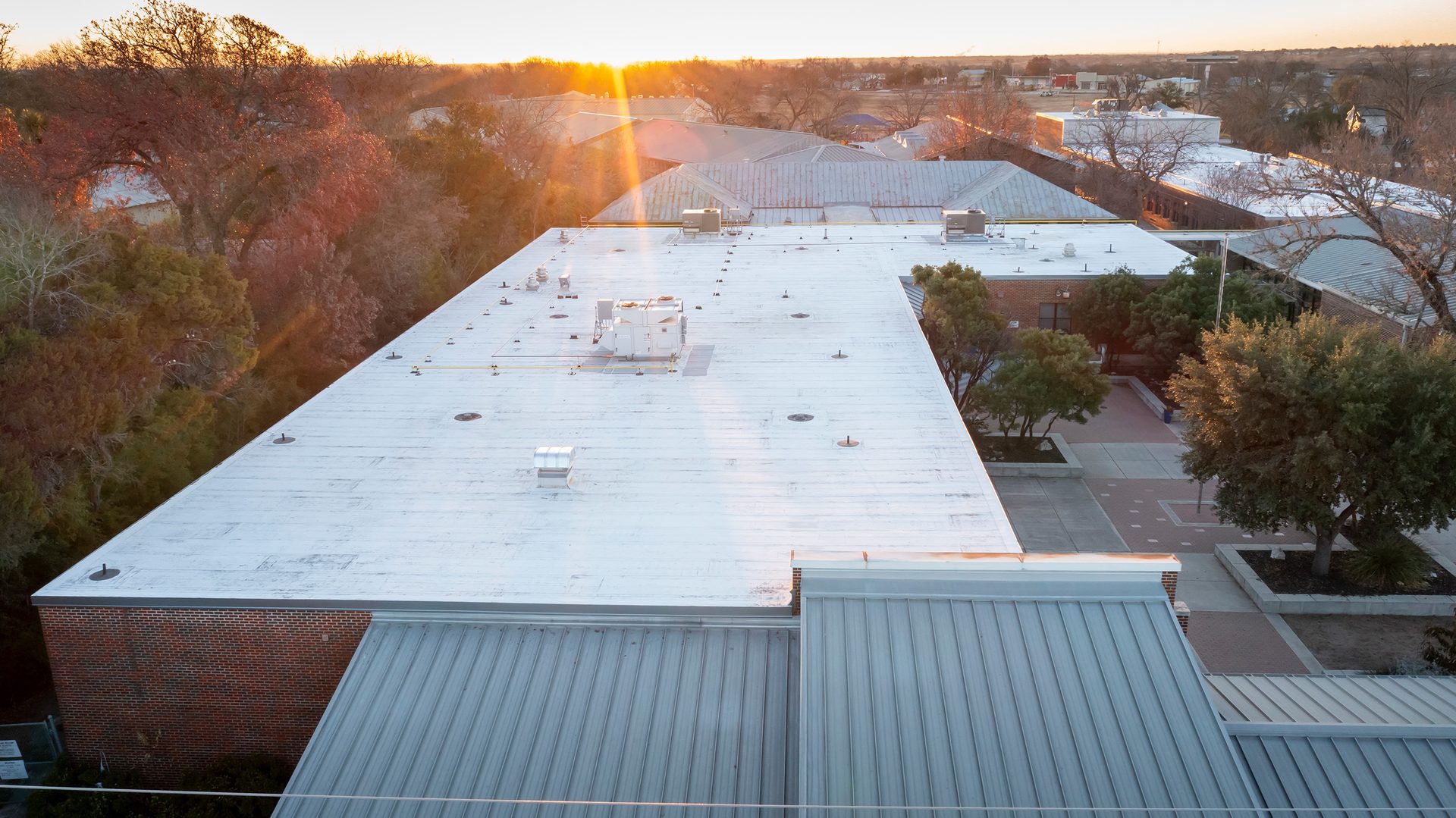 Aerial view of flat roofs and buildings under a golden sunset, surrounded by trees.