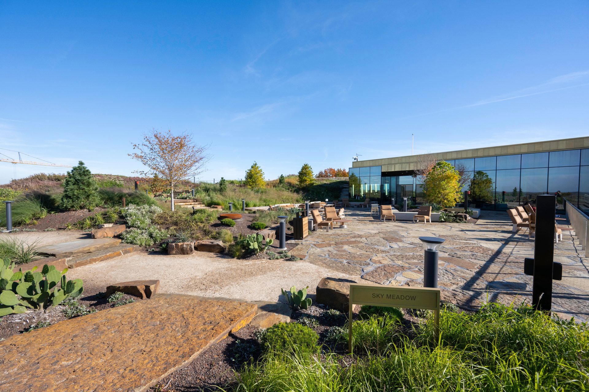 An expansive rooftop garden called 'Sky Meadow' with a patio, seating, and a modern glass building under blue sky.