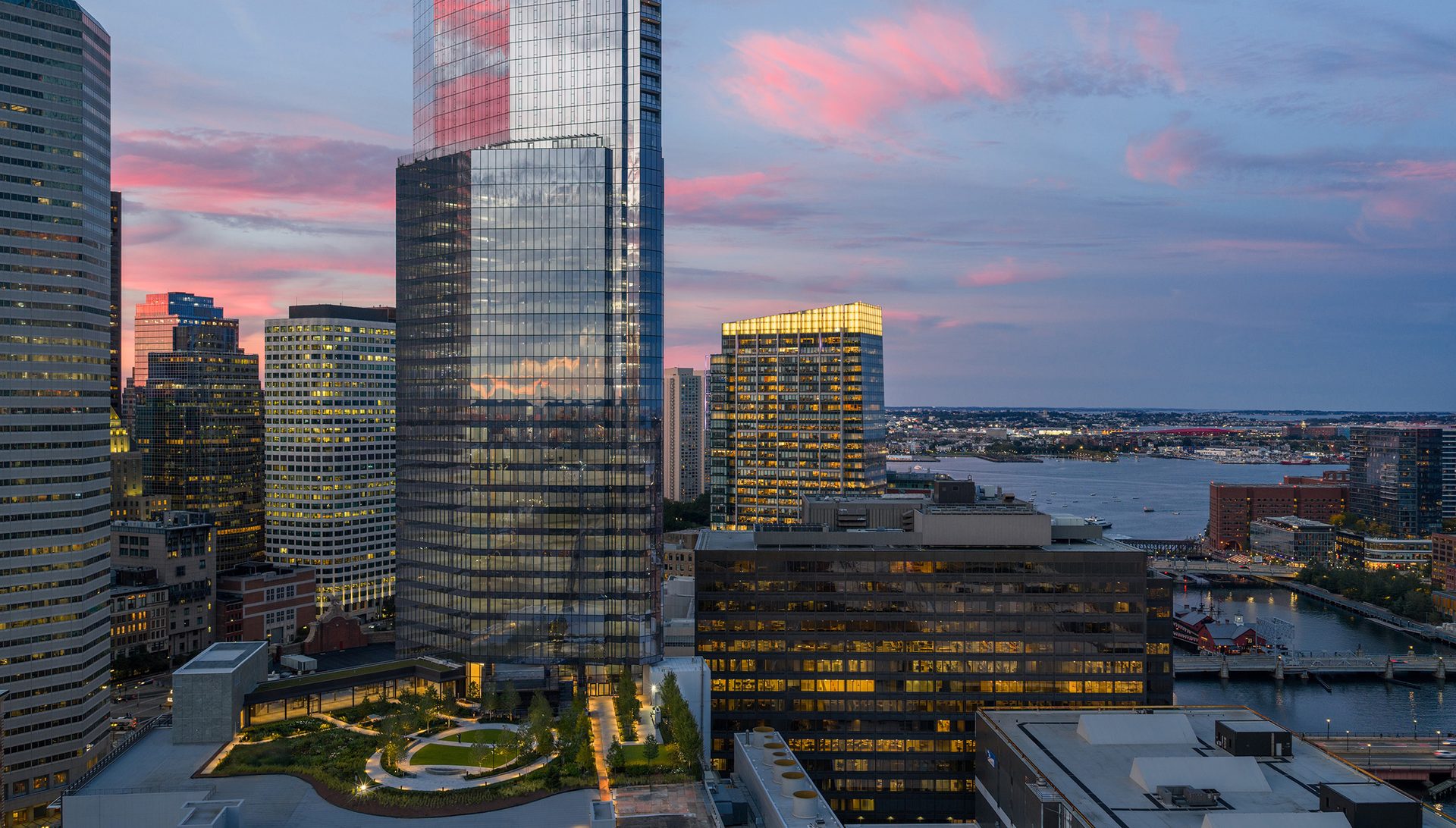 Aerial view of a modern city at dusk with a tall glass skyscraper, rooftop garden, and waterfront.