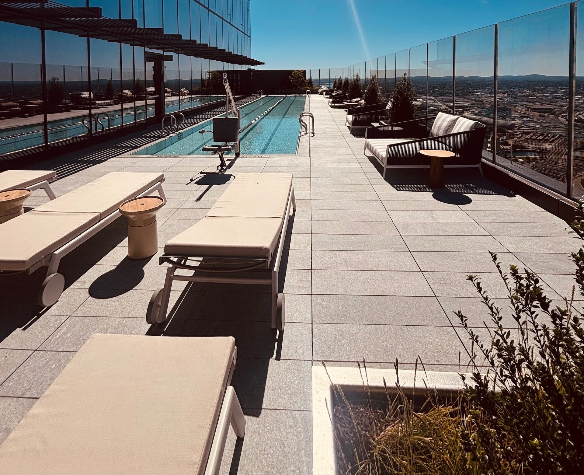A sunny rooftop infinity pool and patio with lounge chairs overlooking a city skyline.