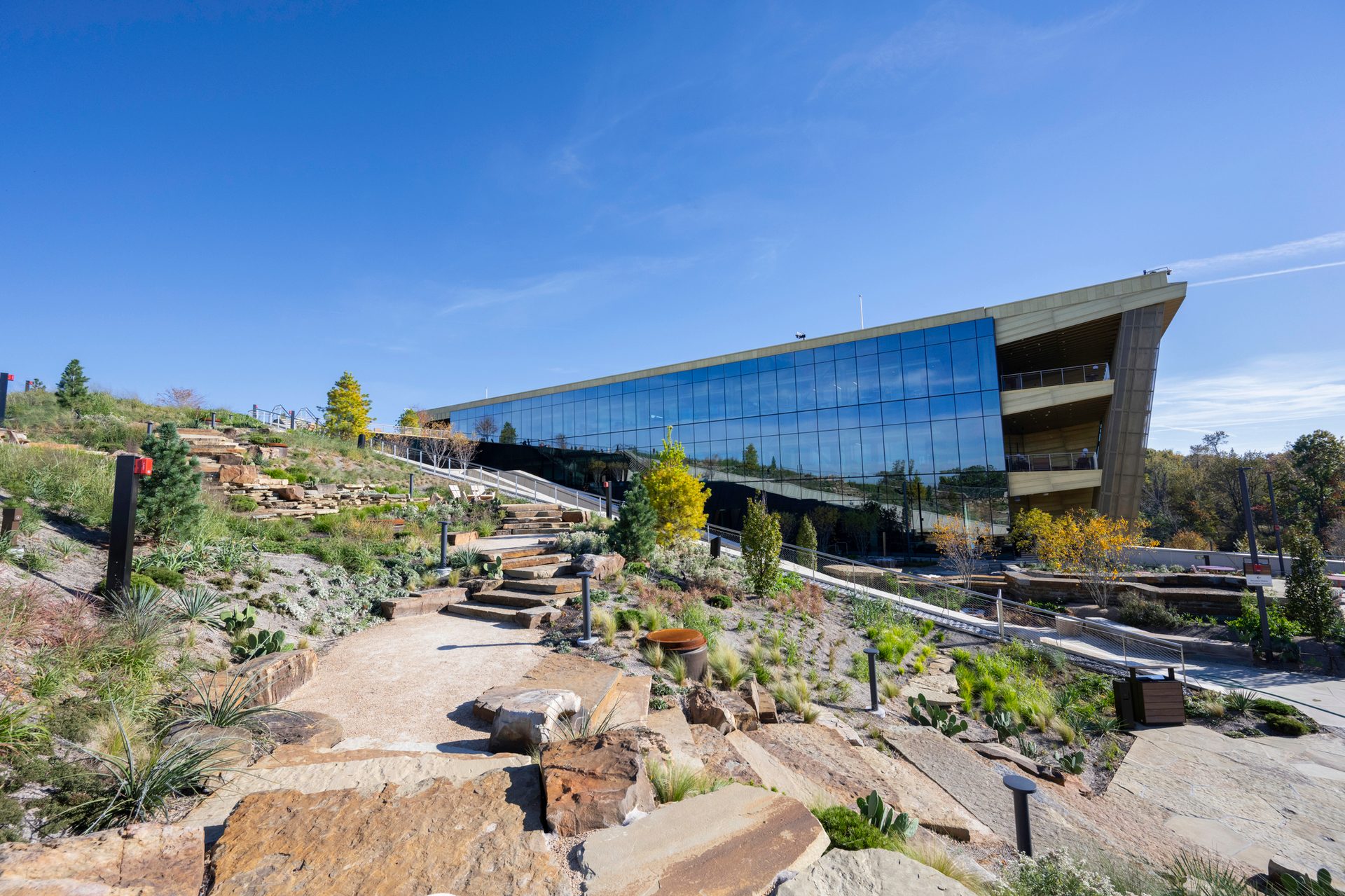Modern glass building seamlessly integrated with terraced rock gardens and stairs under a clear blue sky.